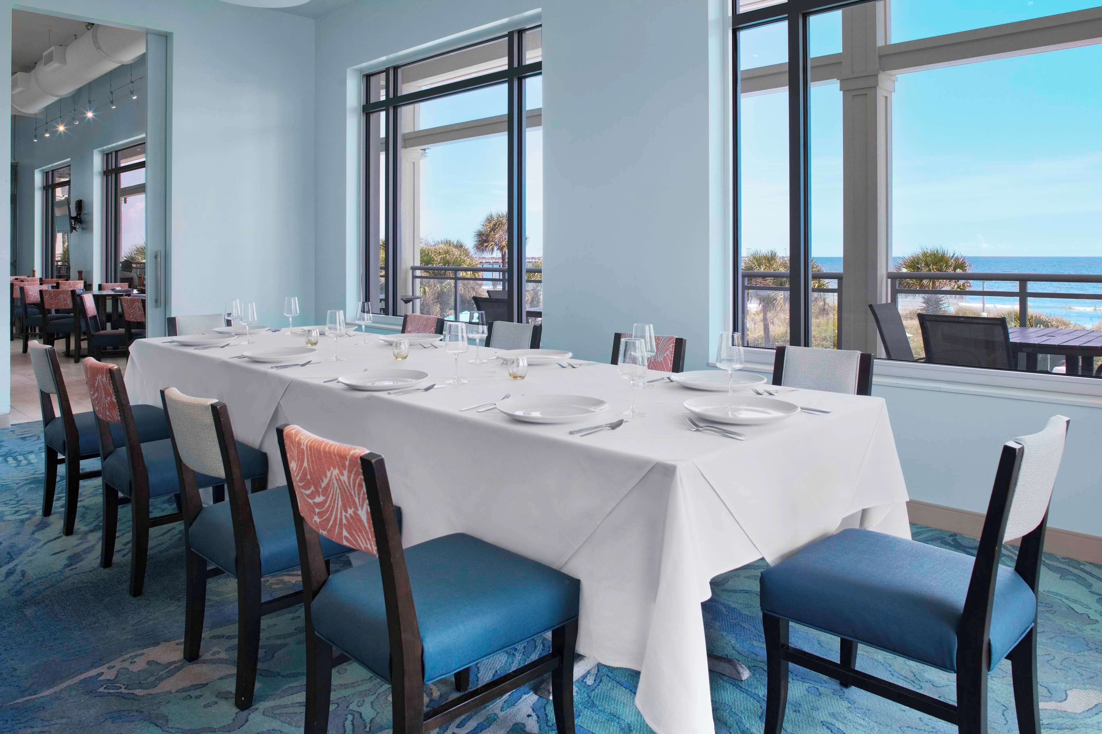 A dining area with a long table set for six, large windows, and a view of the ocean.