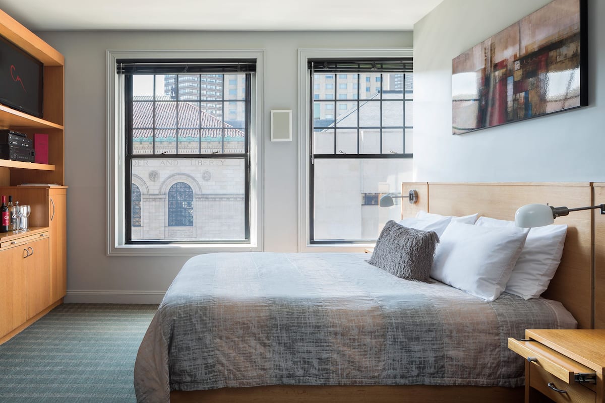 A bedroom with a large bed, two windows, a wooden dresser, and a painting on the wall.
