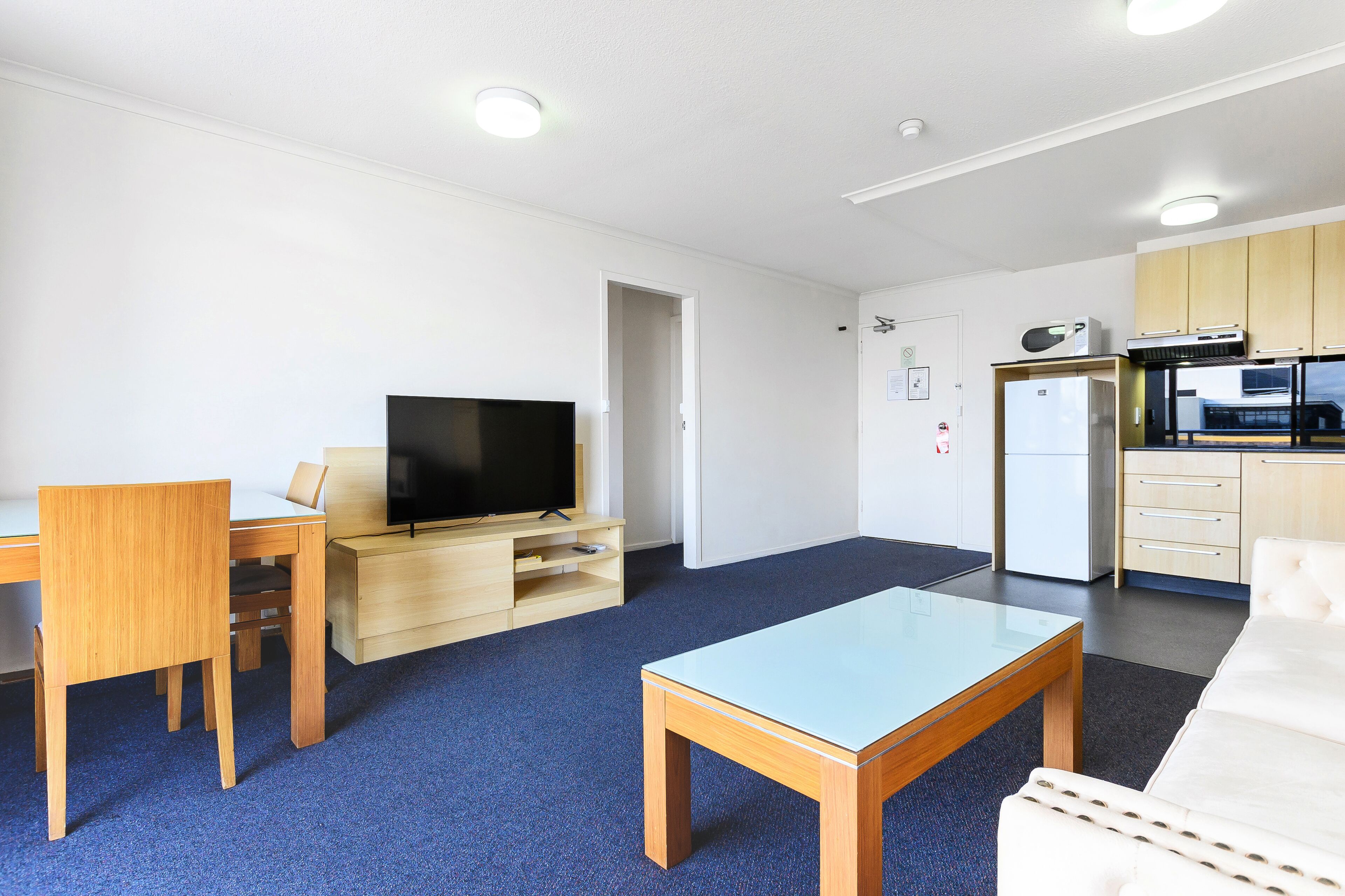 A modern living room with a television, wooden furniture, a glass coffee table, and a kitchenette with a refrigerator and microwave.