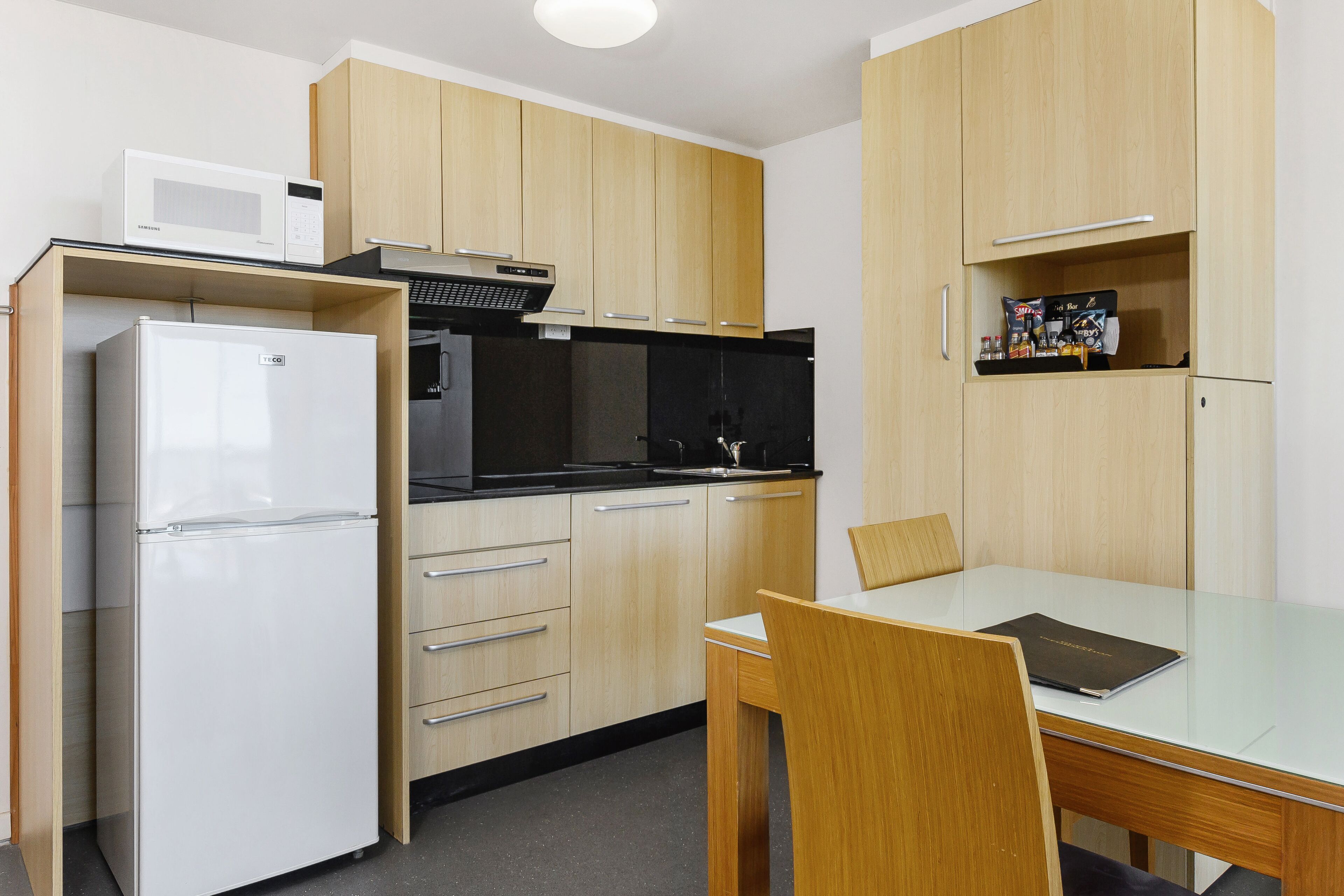 A compact kitchen with a refrigerator, microwave, and black stove top oven. There is a wooden dining table with a chair.