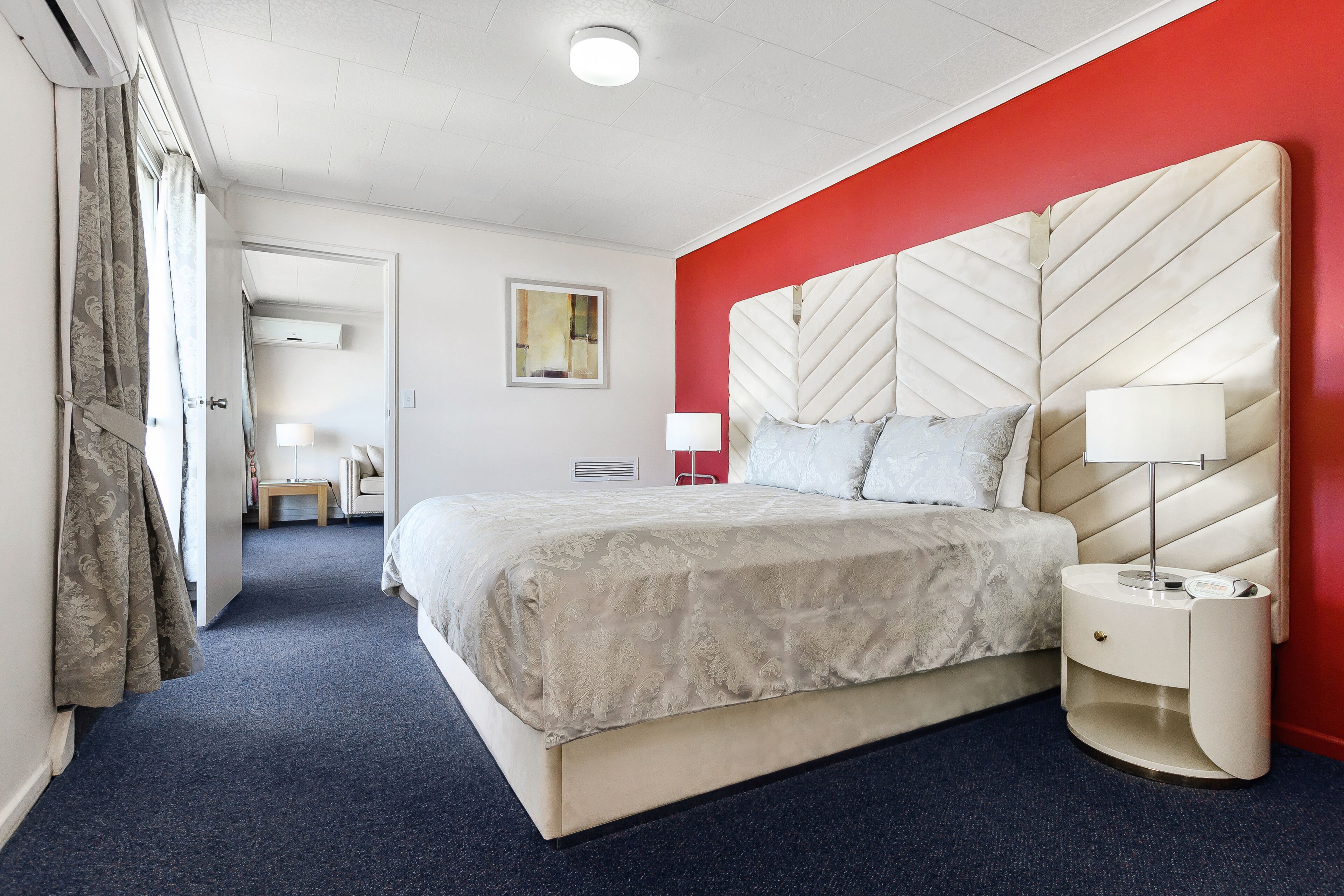 Una habitación de hotel con una cama grande, una mesita de noche, una pared de acento roja, una lámpara en el techo y una puerta que da a otra habitación.