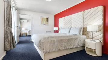 Una habitación de hotel con una cama grande, una mesita de noche, una pared de acento roja, una lámpara en el techo y una puerta que da a otra habitación.