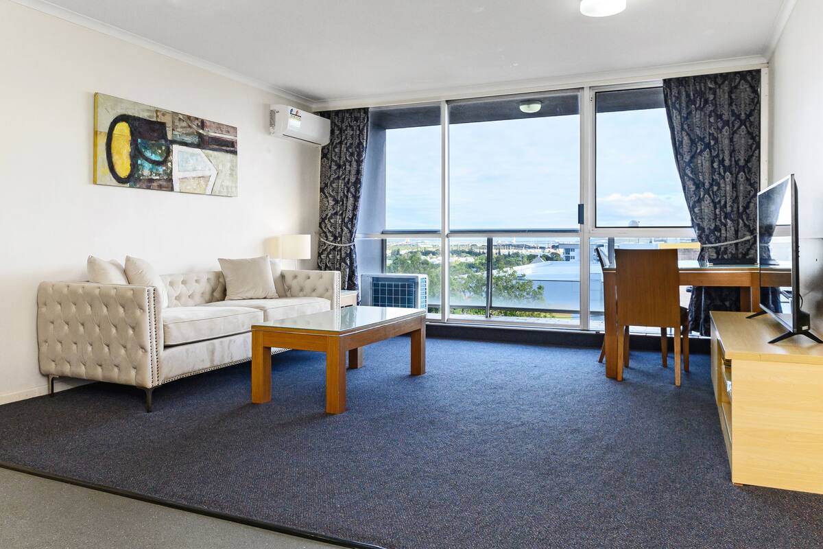 Una sala moderna con sofá, mesa de centro y escritorio con silla. Hay un ventanal con cortinas y un equipo de aire acondicionado en la pared.
