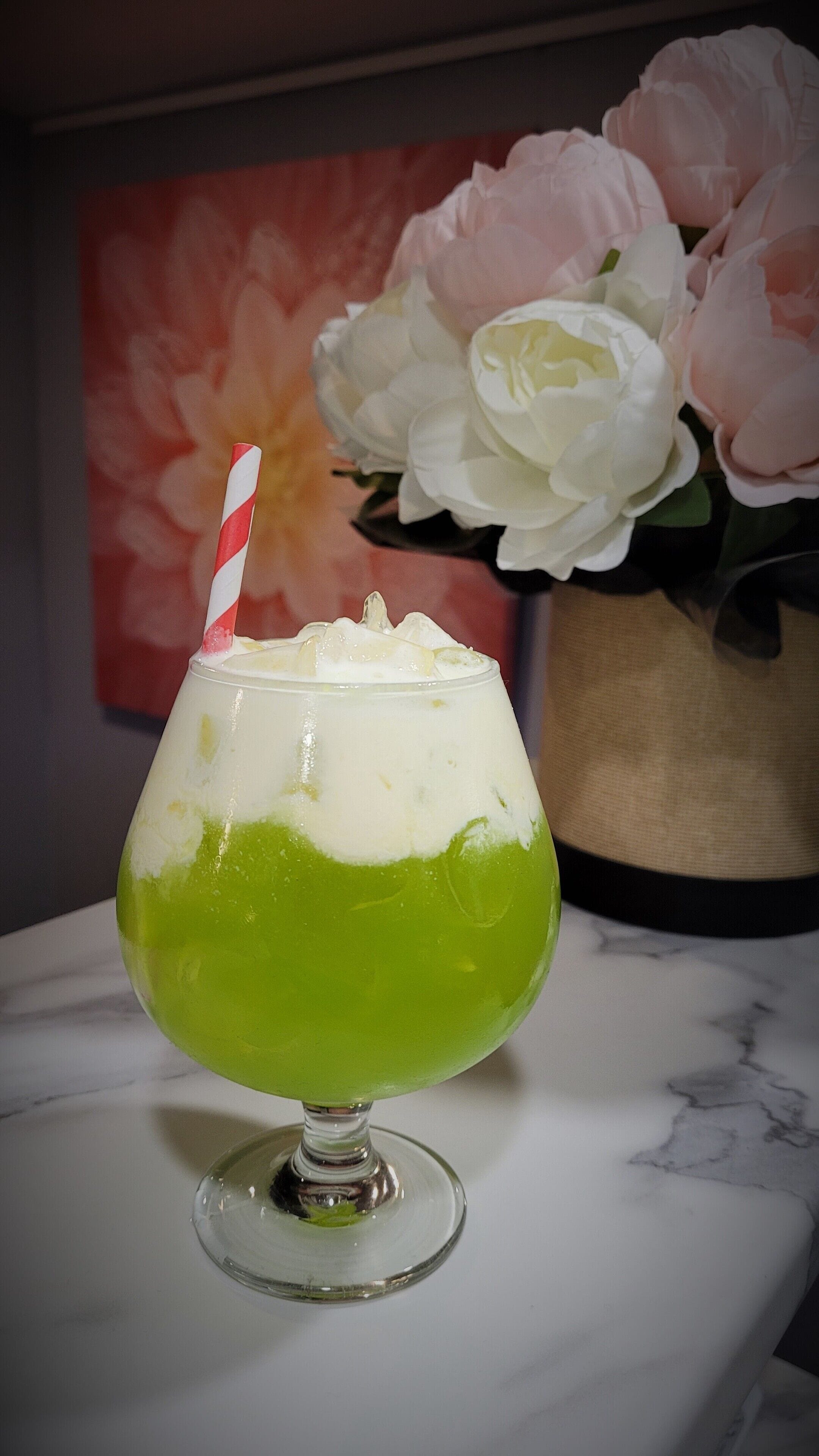 Un cóctel en un vaso con pajita rayada, una capa verde y una capa de espuma blanca, sobre una superficie de mármol.