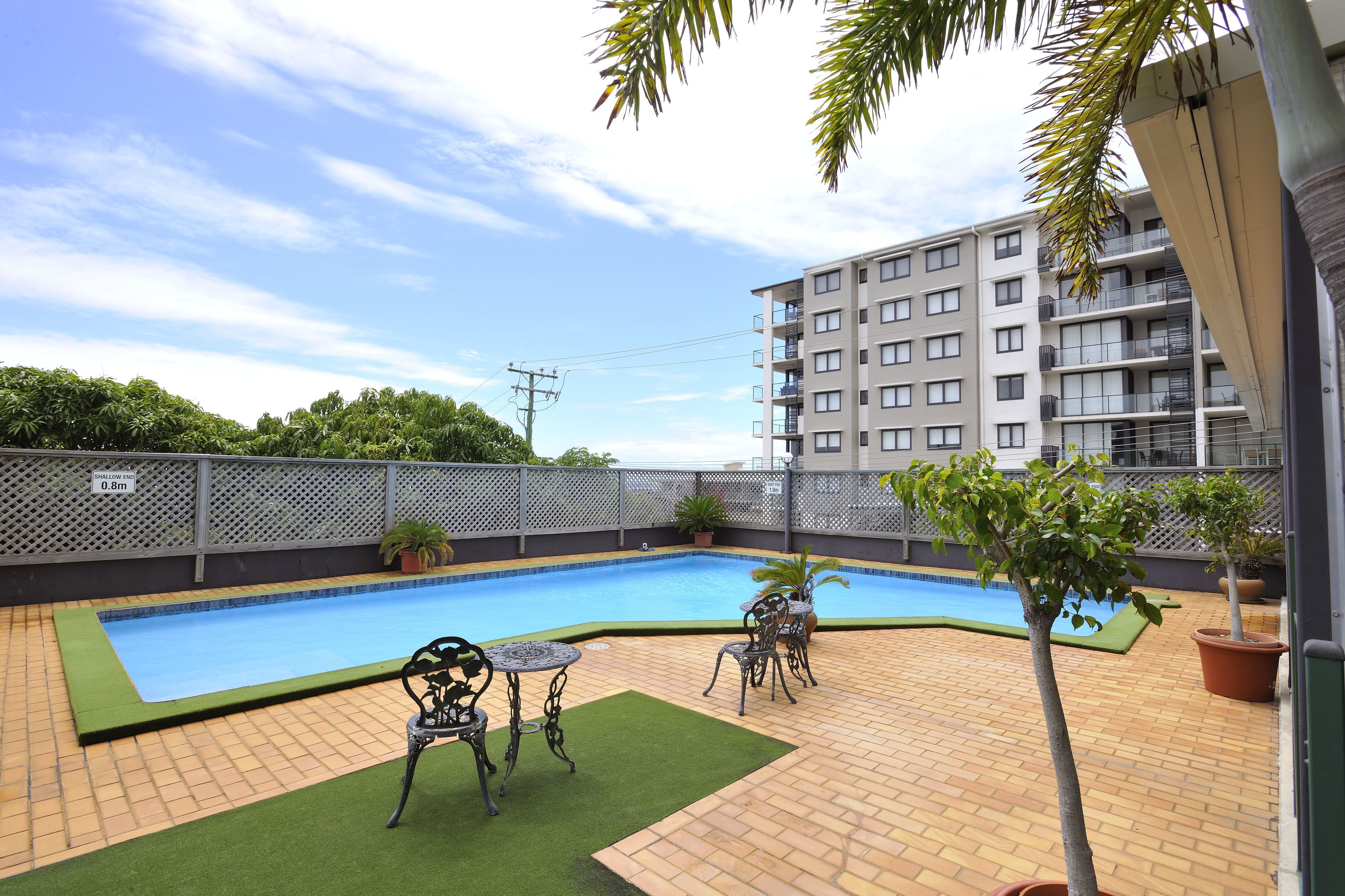 Piscina com deck revestido, cercada por uma grade, e um edifício ao fundo.