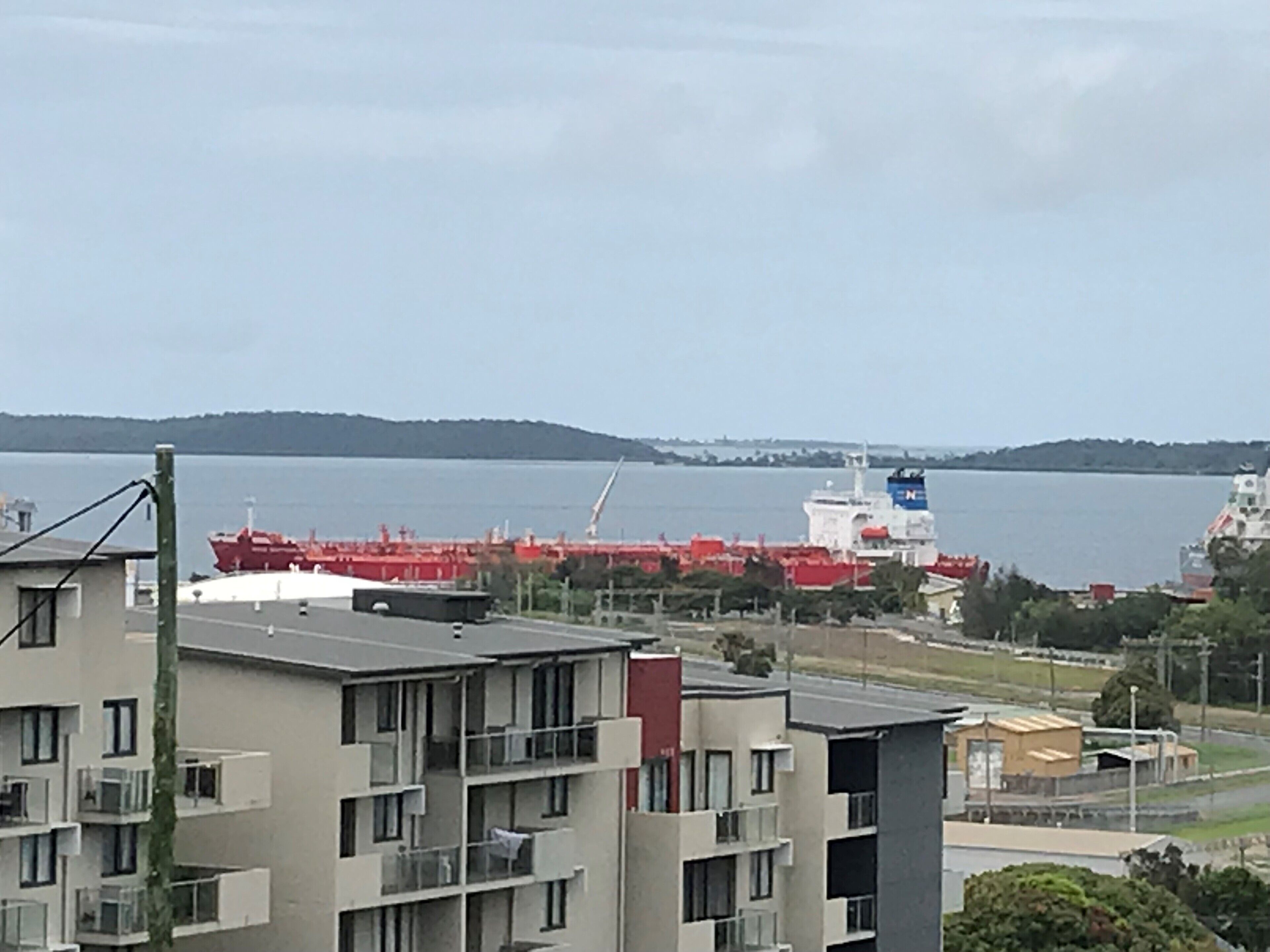 A coastal area with a large red cargo ship docked, a multi-story building with balconies, and a view of the water and distant hills.
