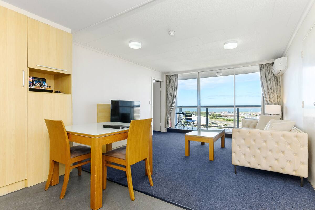 Habitación de hotel con zona de comedor, sofá, televisor y un amplio ventanal con vista al mar.