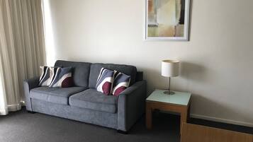 Habitació d'hotel moderna amb un sofà gris, dos coixins decoratius, una petita taula auxiliar amb una làmpada i un quadre abstracte emmarcat a la paret.