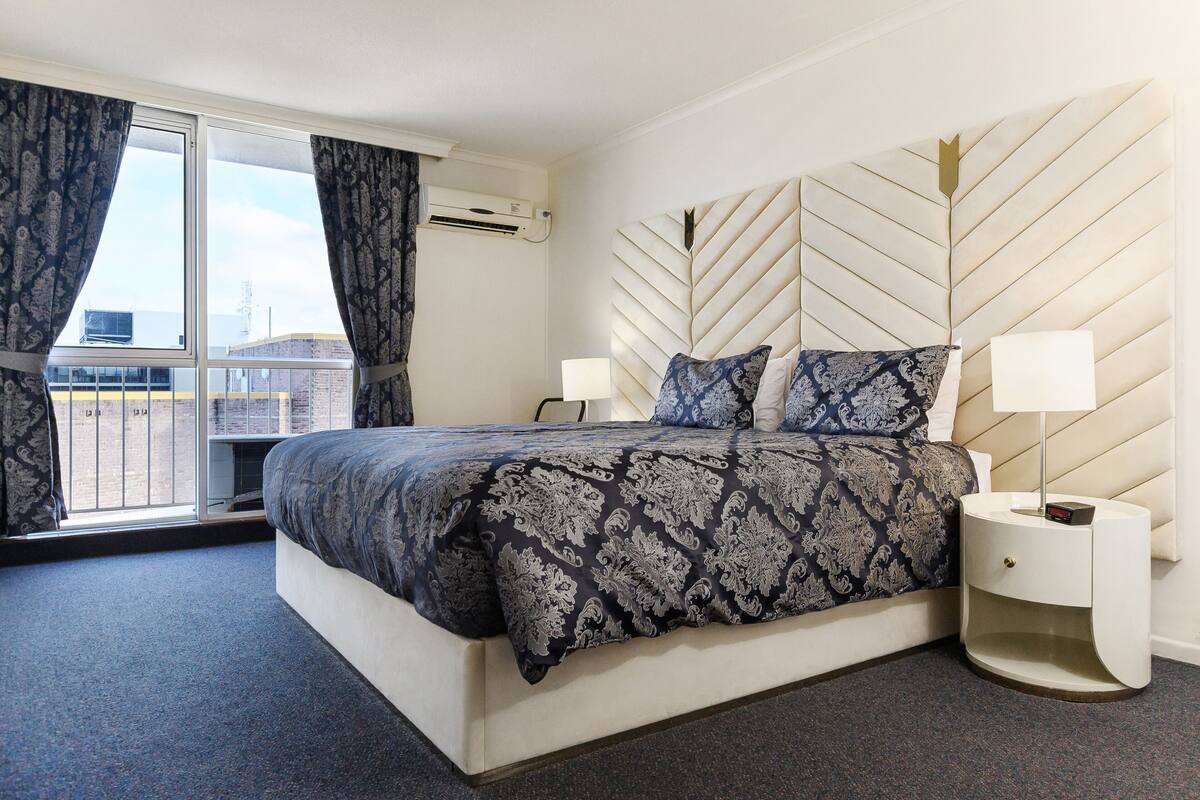 Un dormitorio con cama, mesitas de noche, una ventana con cortinas y un cabecero estampado con hojas.