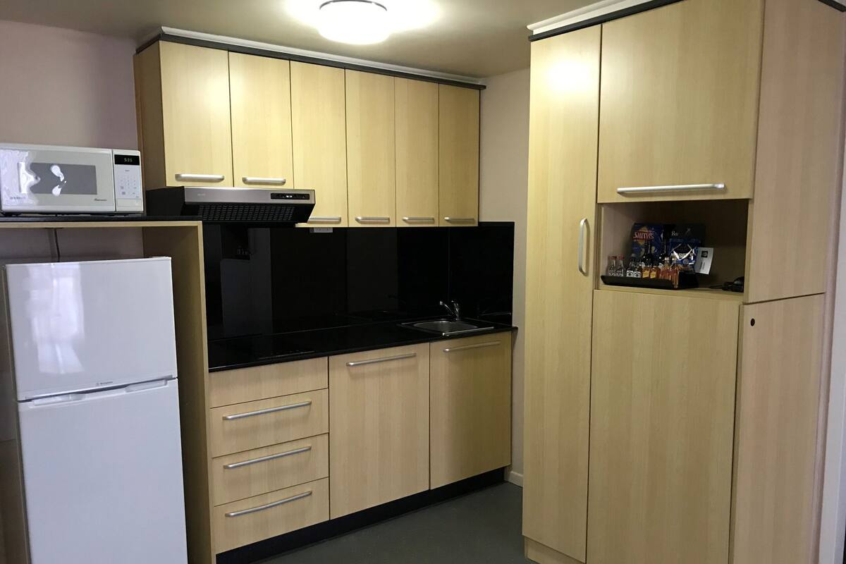 Una cocina compacta con armarios de madera clara, encimera negra, refrigerador blanco y microondas.