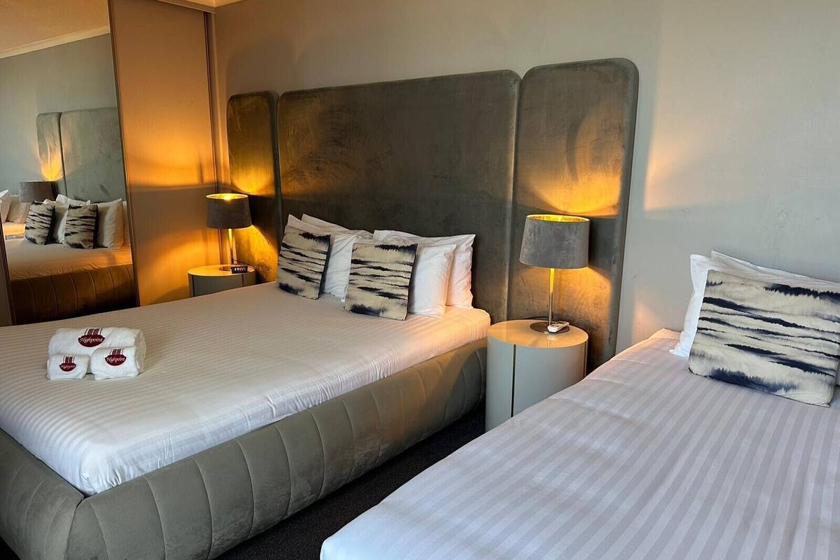 Habitación de hotel con dos camas, cada una con cabecero, lámparas de noche y almohadas. Hay toallas sobre las camas y un espejo en la pared.