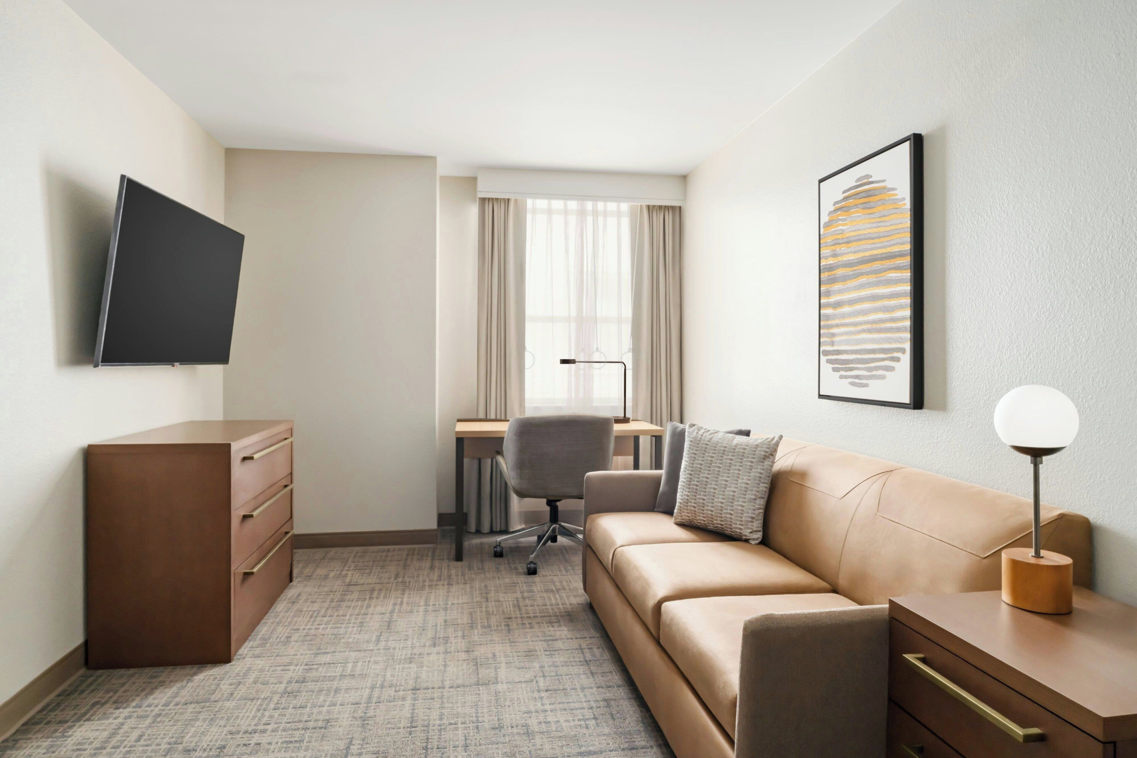 A hotel room with a flat-screen TV, a desk with a chair, a sofa, a bedside table with a lamp, and a framed picture on the wall.