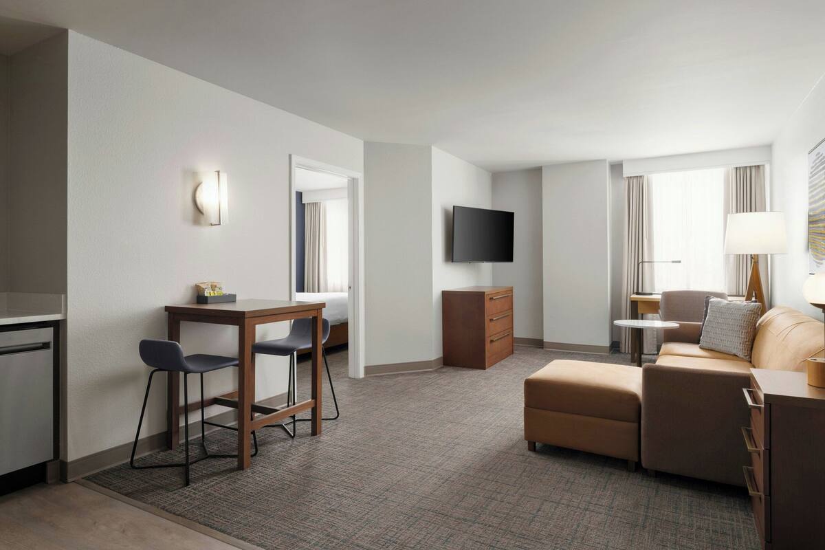 A modern hotel room with a kitchenette, a flat-screen TV mounted on the wall, a sofa, a small table, and a lamp.