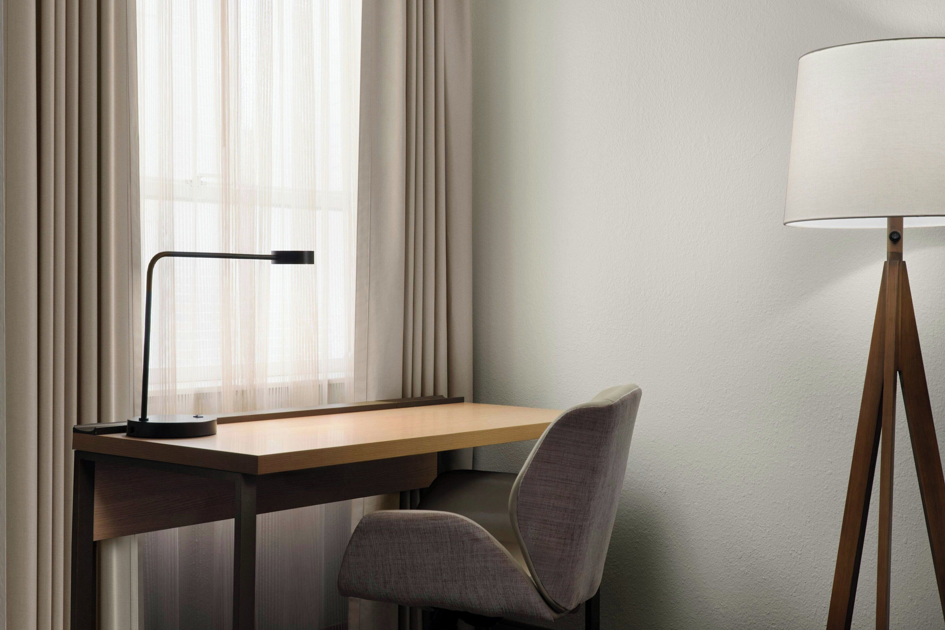 Una habitación de hotel moderna con un escritorio de madera, una silla gris, una lámpara de pie y cortinas de gasa.