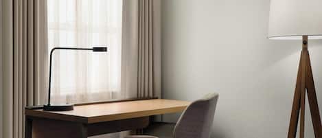 Una habitación de hotel moderna con un escritorio de madera, una silla gris, una lámpara de pie y cortinas de gasa.