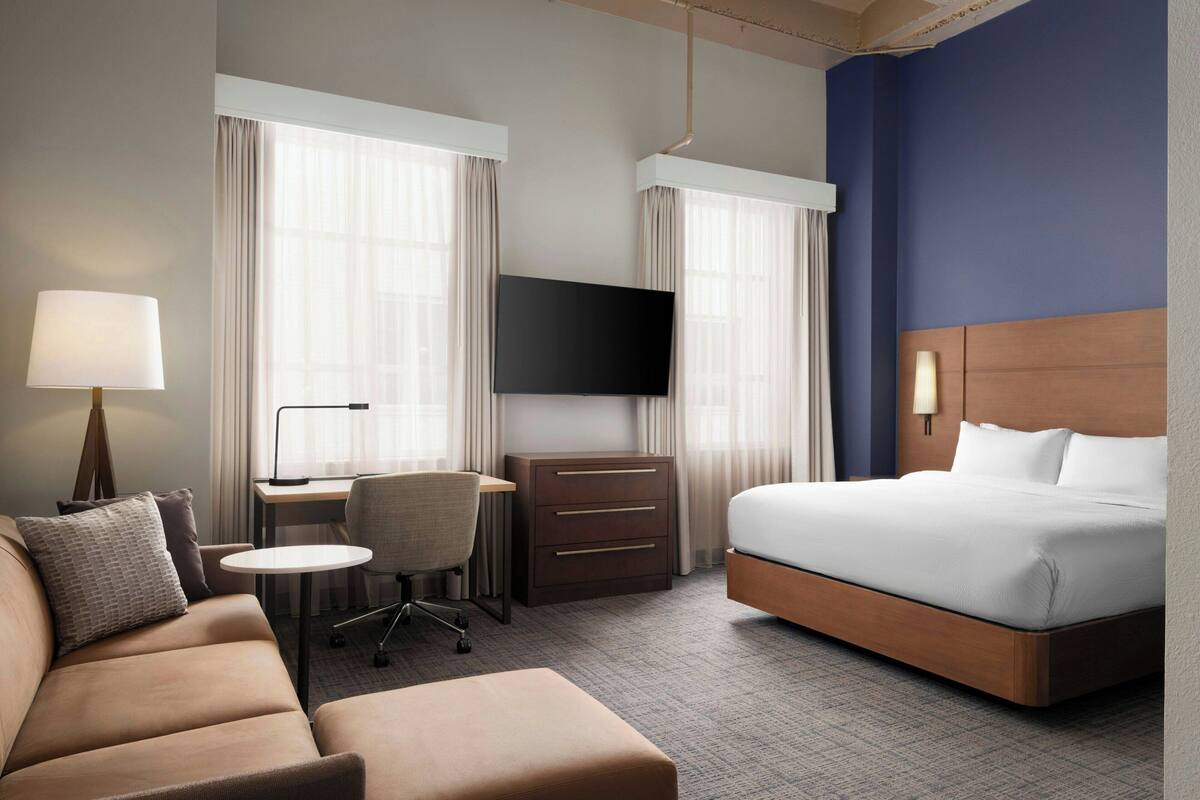 A hotel room with a bed, a sofa, a desk with a chair, a TV, and a lamp.
