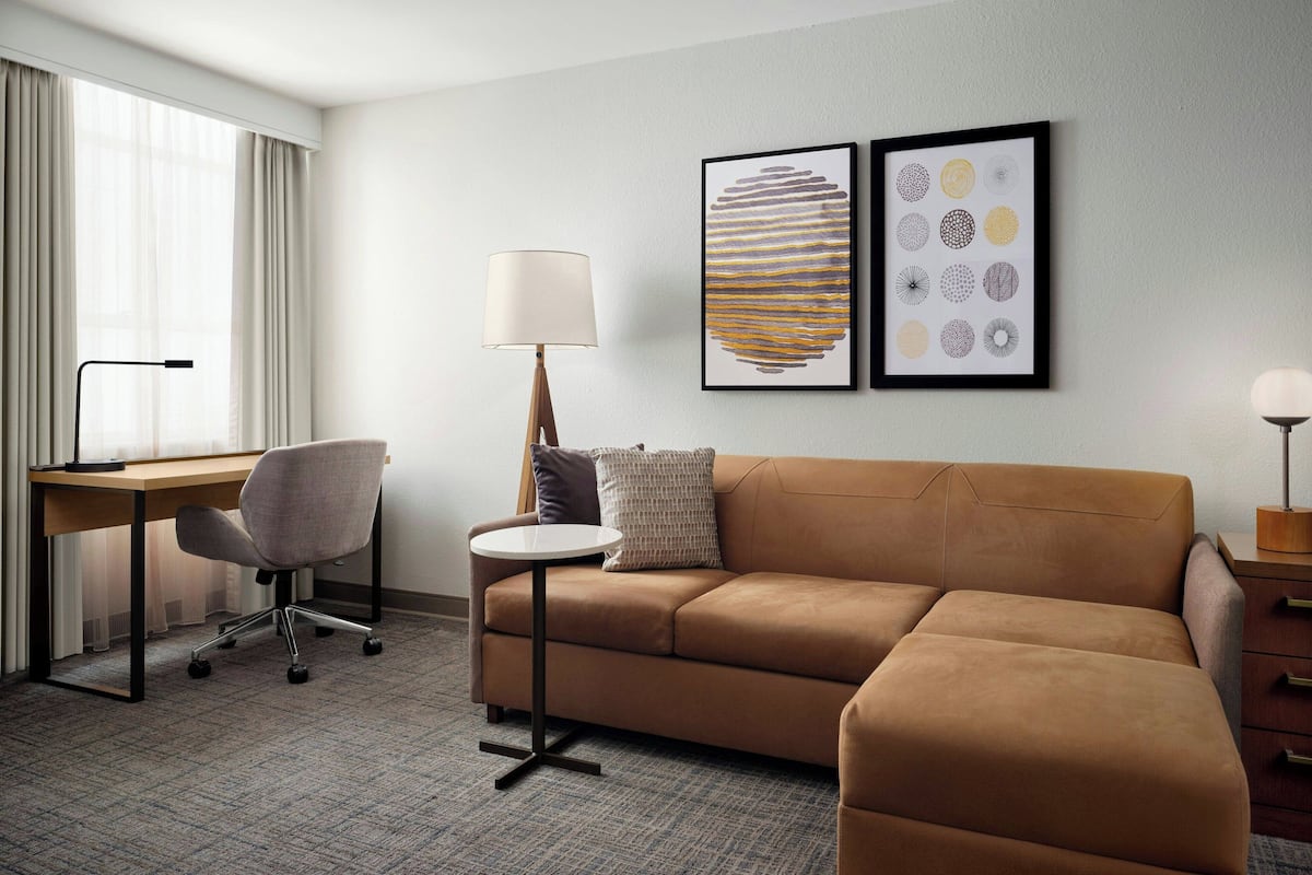 A modern hotel room with a brown sofa, a desk, a chair, and two framed artworks on the wall.