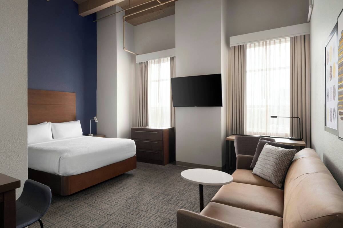 A hotel room with a bed, a sofa, a desk, a TV, and a window with curtains.