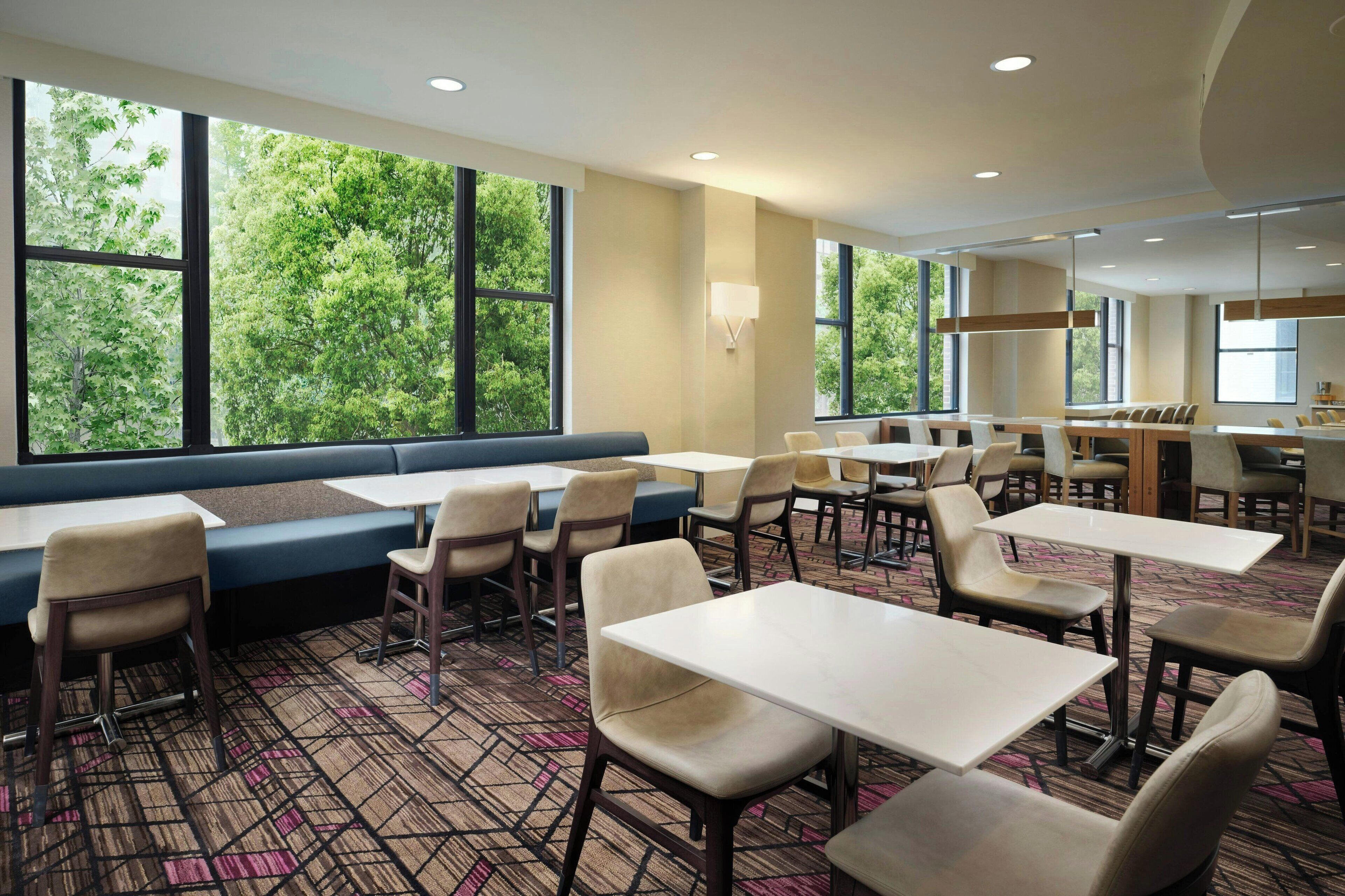 Un espace de restauration d’hôtel avec des tables, des chaises, un banc et de grandes fenêtres donnant sur la verdure.
