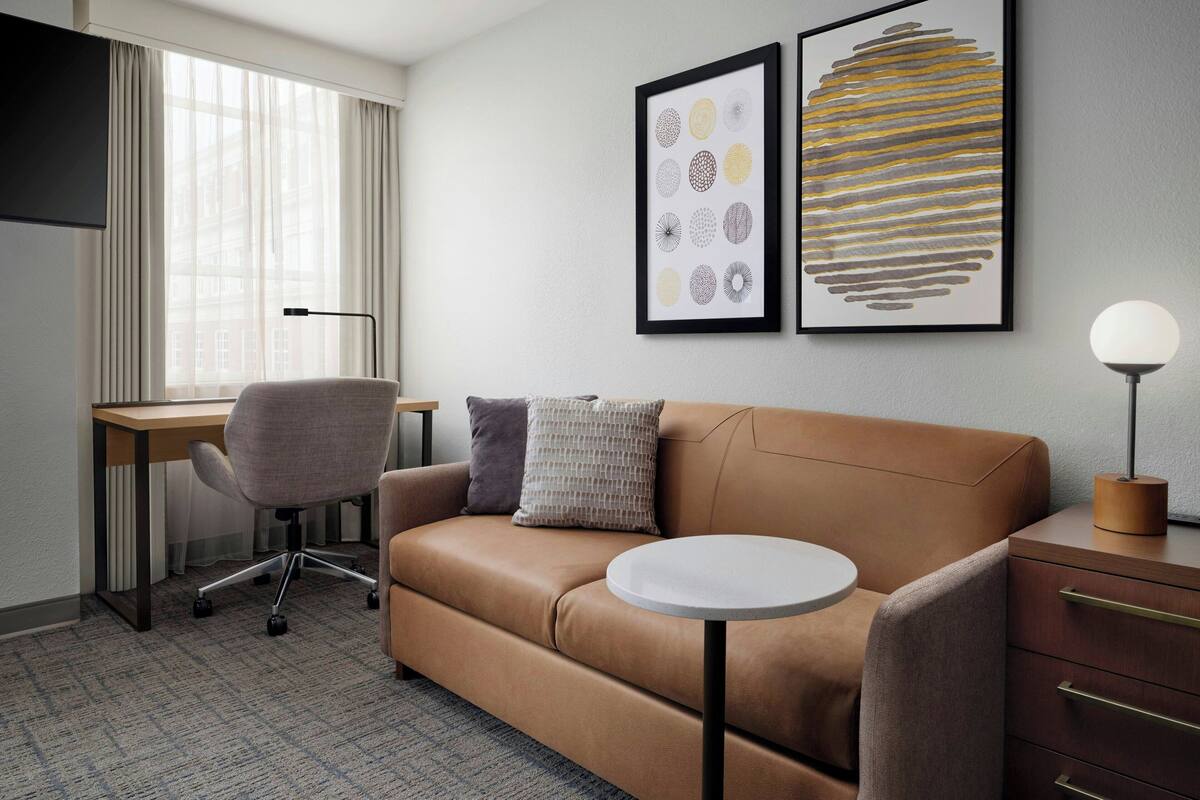 A modern hotel room with a leather sofa, a desk, a chair, a round table, and two framed artworks on the wall.
