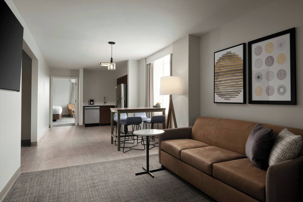 A modern living room with a brown sofa, a round coffee table, and a dining area with a table and chairs. There are framed artworks on the wall and a lamp on the sofa.
