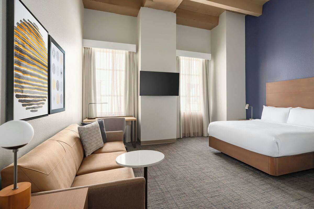 A hotel room with a bed, a sofa, a small table, a TV, and a window with curtains.