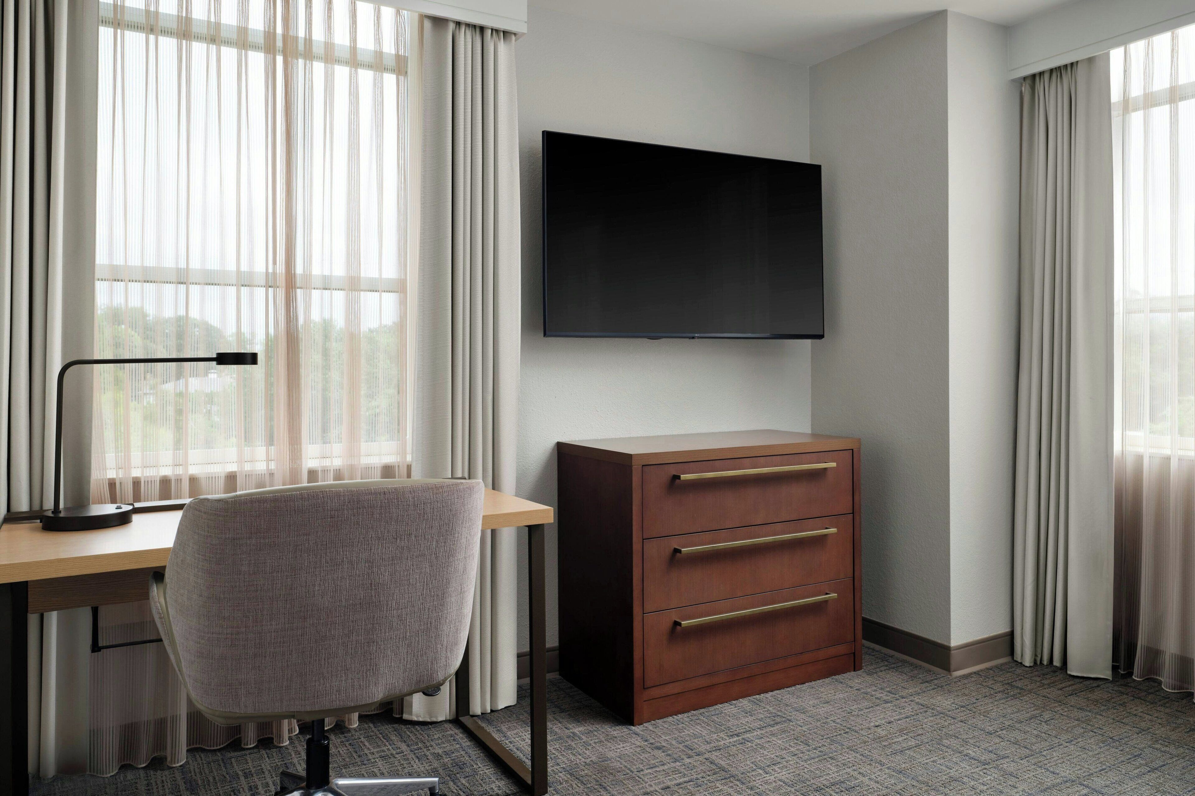 A hotel room with a desk, chair, TV, and a wooden dresser.