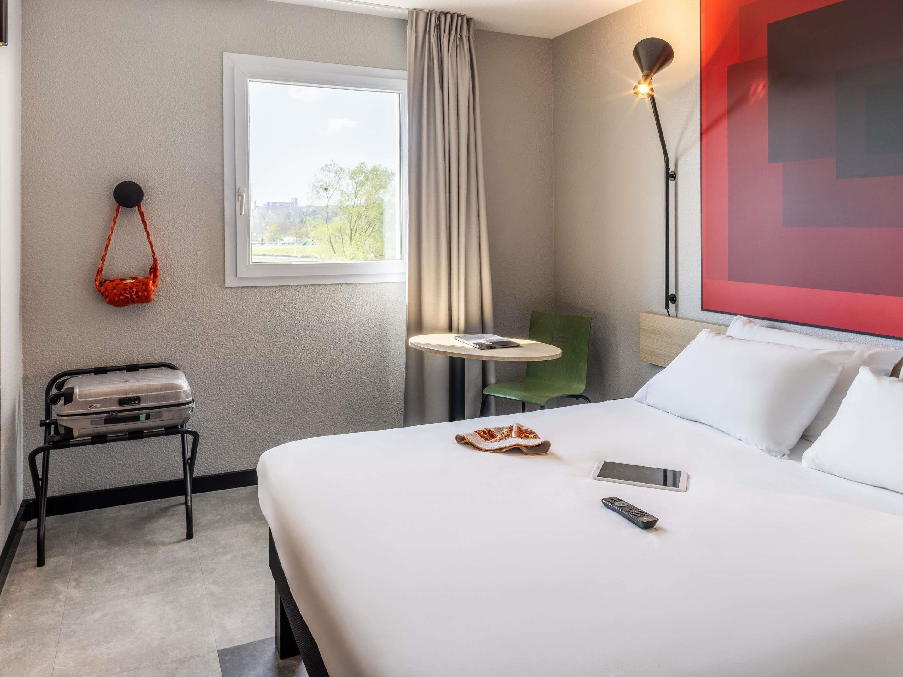 Habitación de hotel con cama, escritorio, silla, ventana, lámpara y maleta.