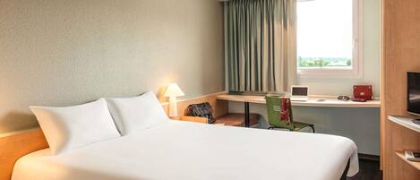 A hotel room with a large bed, a desk with a laptop, a chair, a TV, and a window with curtains.