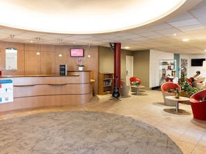 A reception area with a curved wooden desk, a flat-screen TV, a red pillar, and a seating area with red chairs and a small table. - ibis Auch (Auch)