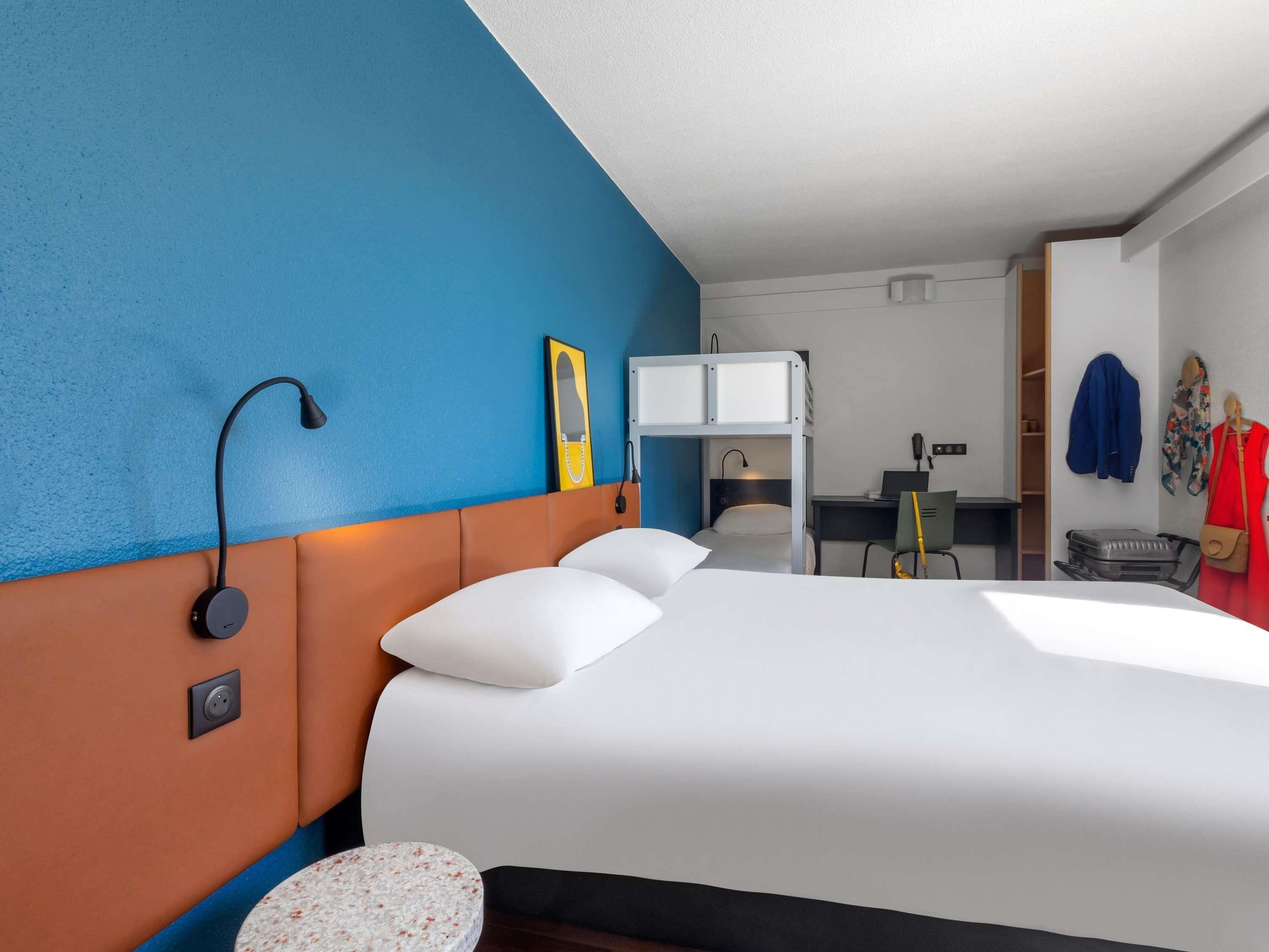 Habitación de hotel con literas, un escritorio con silla, una maleta y un perchero con chaquetas.