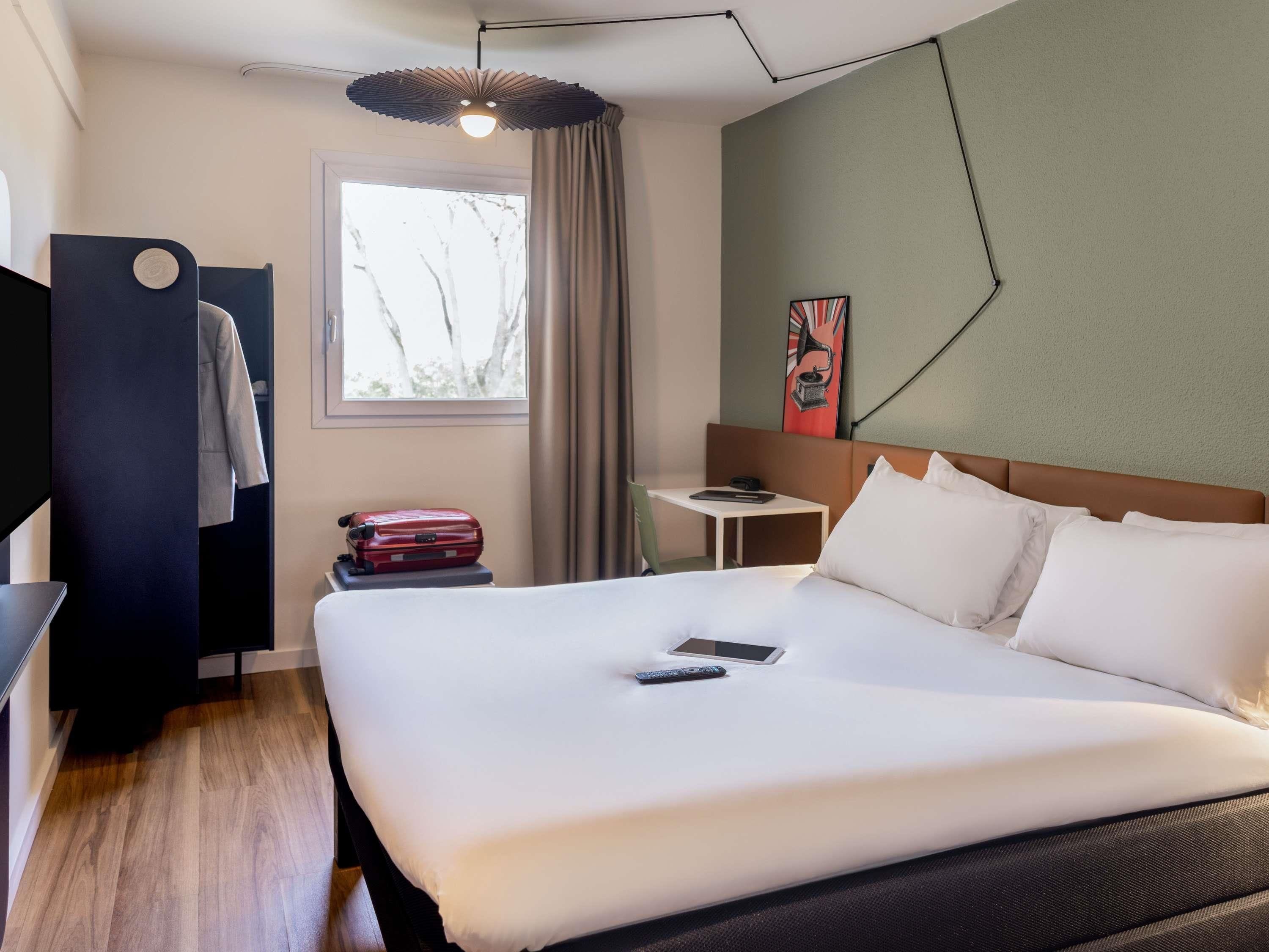 Habitación de hotel con cama, escritorio, silla, ventana con cortinas, una obra de arte en la pared y una lámpara colgante.