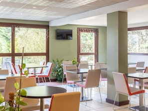 Um restaurante com várias mesas e cadeiras, janelas amplas e uma televisão de tela plana na parede.