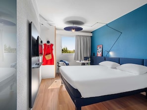 A modern hotel room with a bed, a desk, a chair, a TV, a window with curtains, and a blue accent wall. - ibis Auch (Auch)