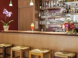Uma área de bar com banquetas, uma bebida e uma estante com garrafas e copos.