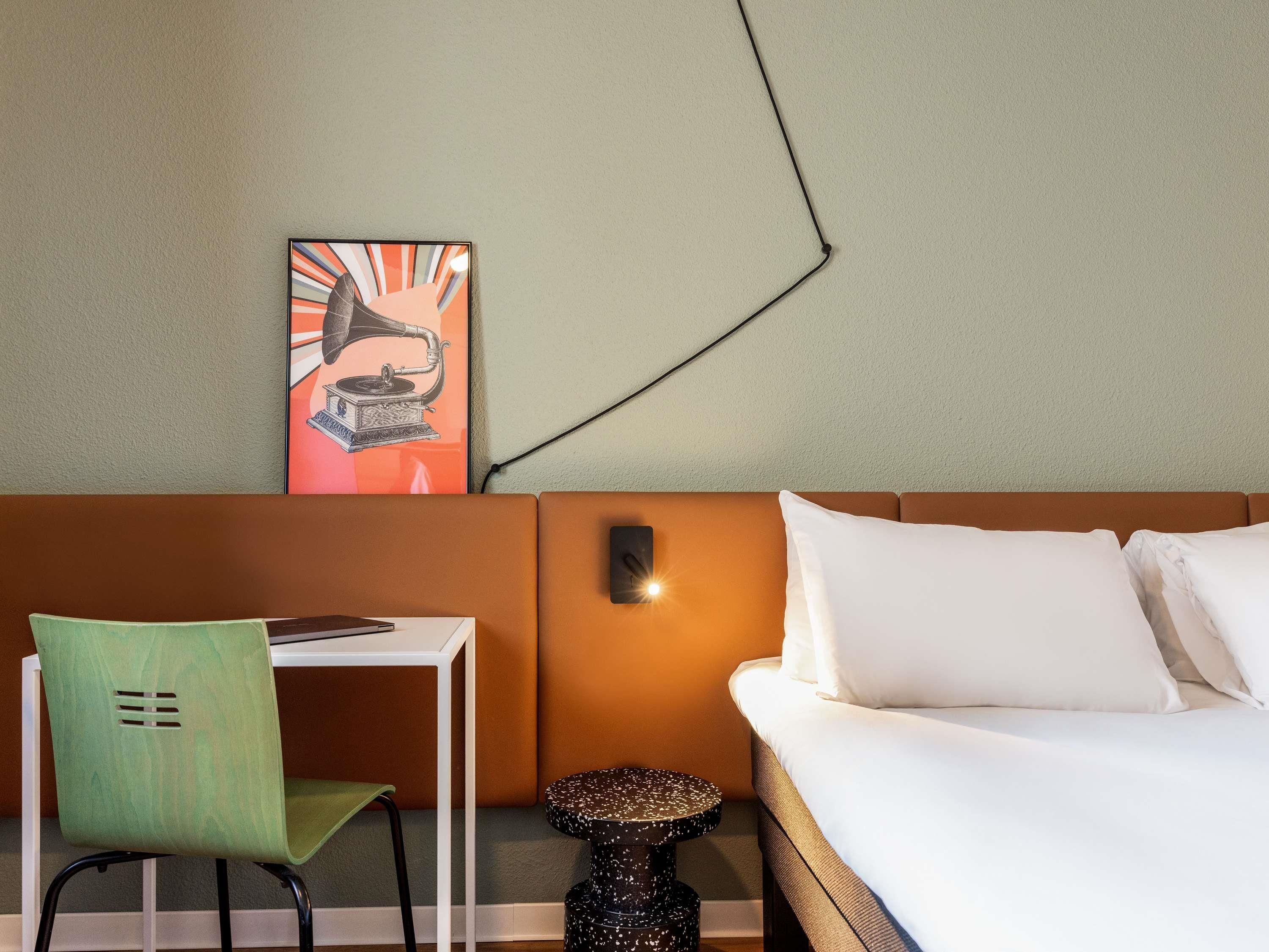 Una habitación de hotel con una cama, una silla verde, una mesita pequeña y una lámpara de pared.