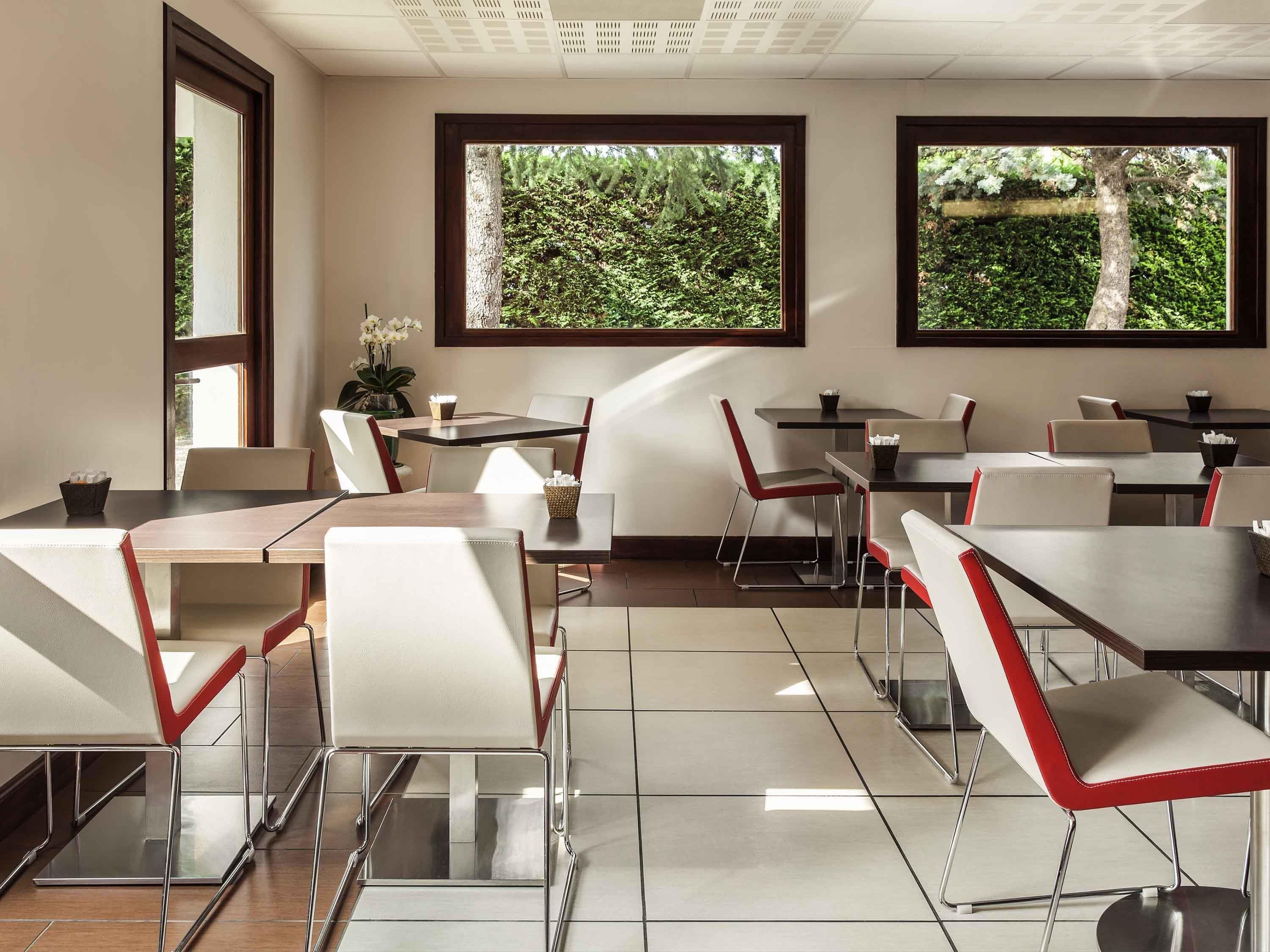 A modern dining area with multiple tables and chairs, large windows with greenery views, and a clean, contemporary design.