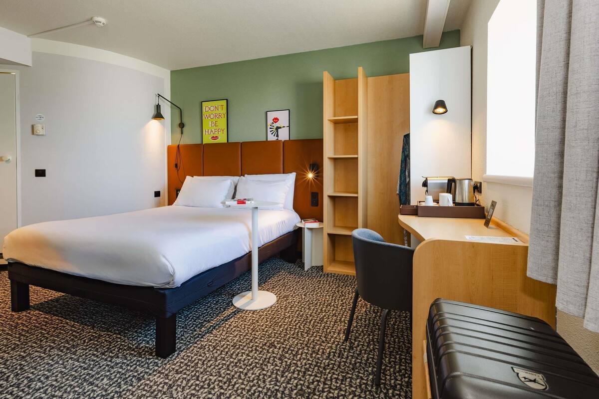 Quarto de hotel com uma cama, uma escrivaninha, uma cadeira, uma mala, um espelho, uma prateleira e uma parede com um quadro e uma placa.