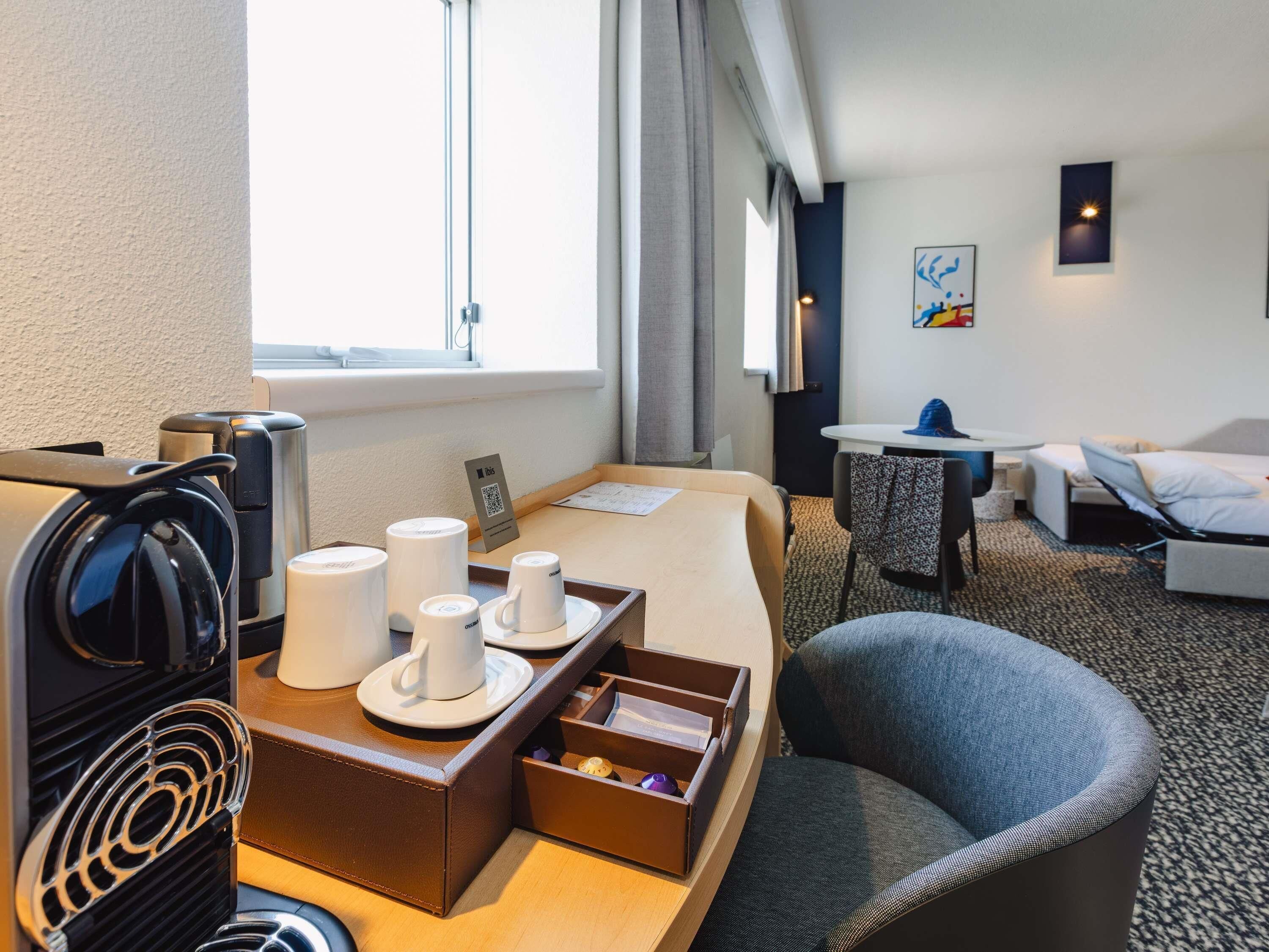 A hotel room with a bed, a desk with a coffee maker, a chair, and a small table with a laptop.