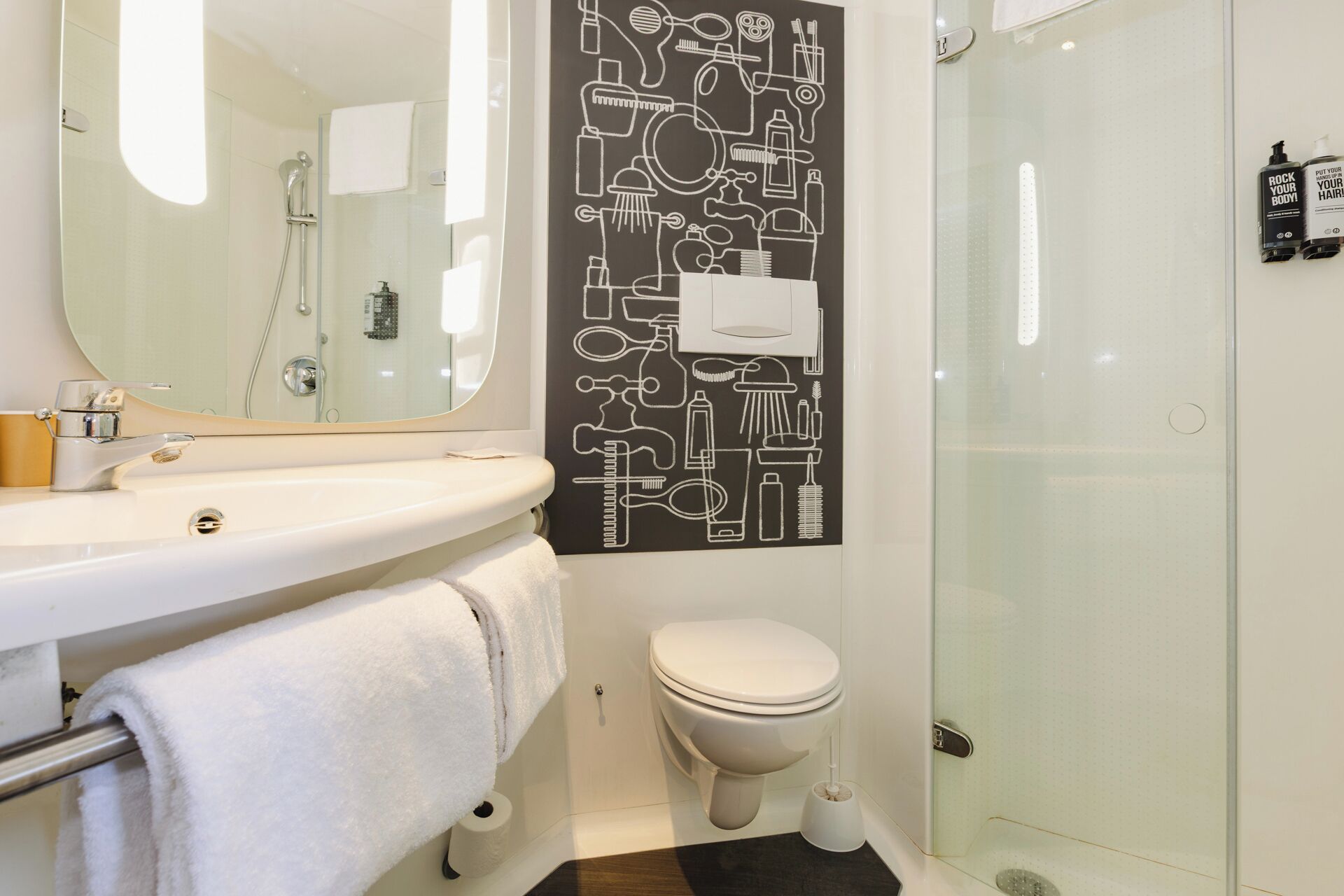 A modern bathroom with a white sink, mirror, and toilet. There is a shower area with a glass door. A black wall decal with bathroom icons is visible.
