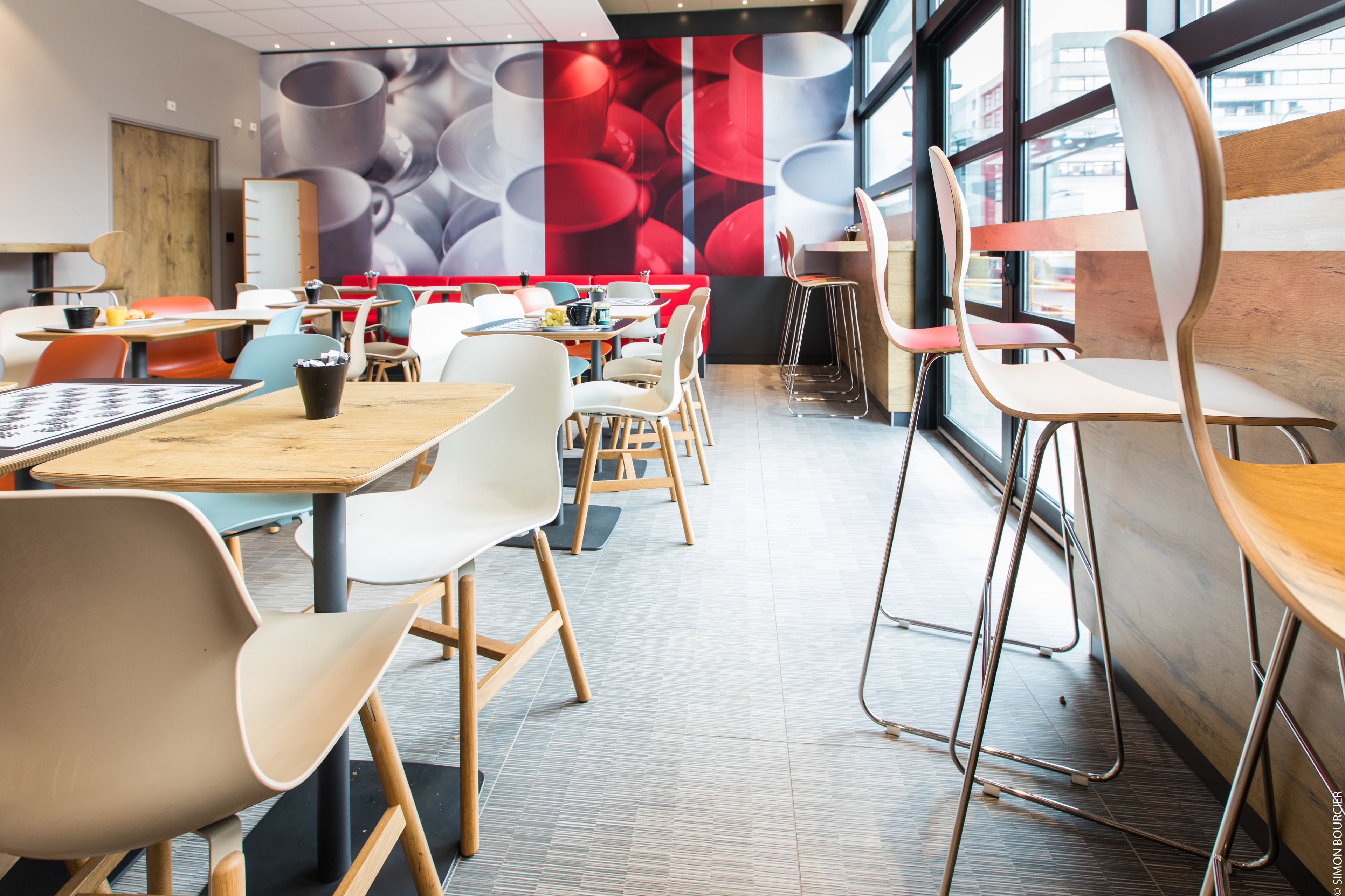Una cafetería moderna con mesas y sillas de madera, un mural grande con tazas de café y ventanas amplias que dejan entrar luz natural.