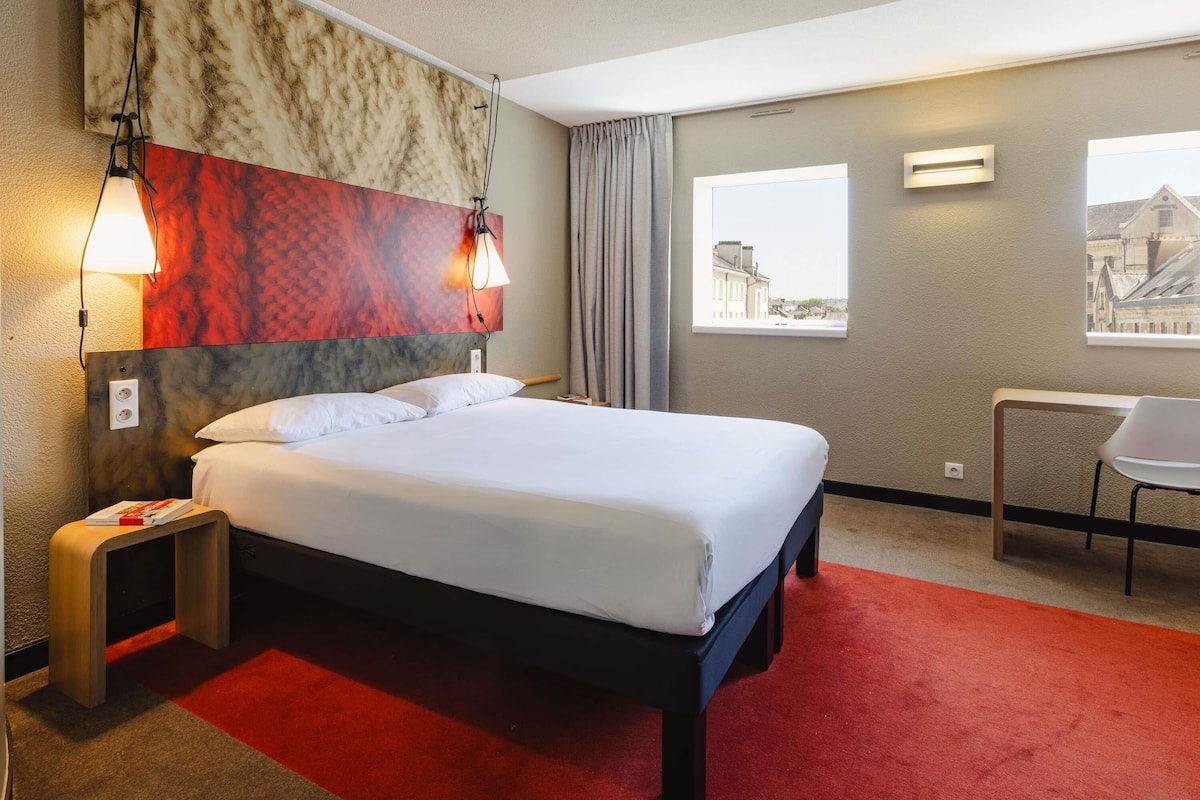 Quarto de hotel com cama, escrivaninha, cadeira e janela com vista para a cidade.