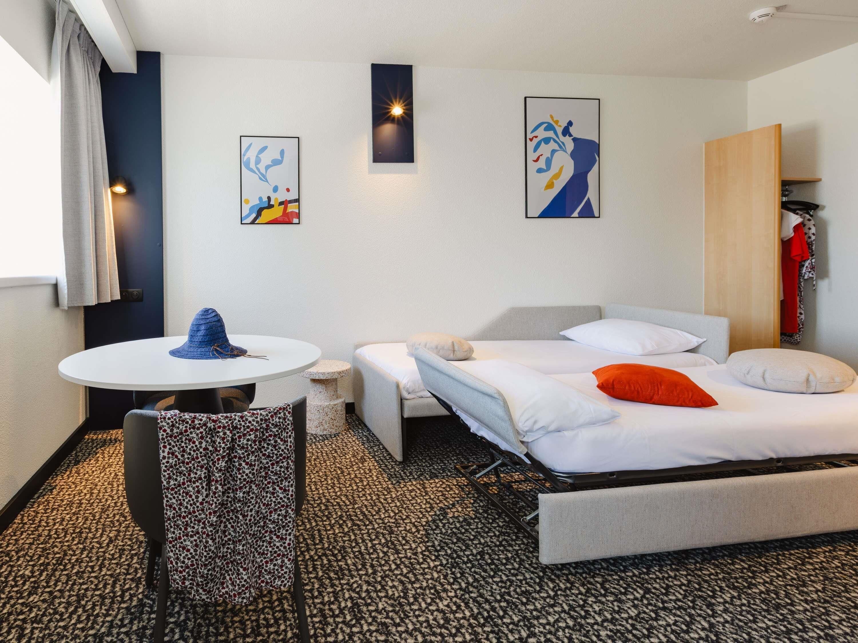 Et hotelværelse med seng, et lille bord, en stol, et skrivebord og billedkunst på væggene.
