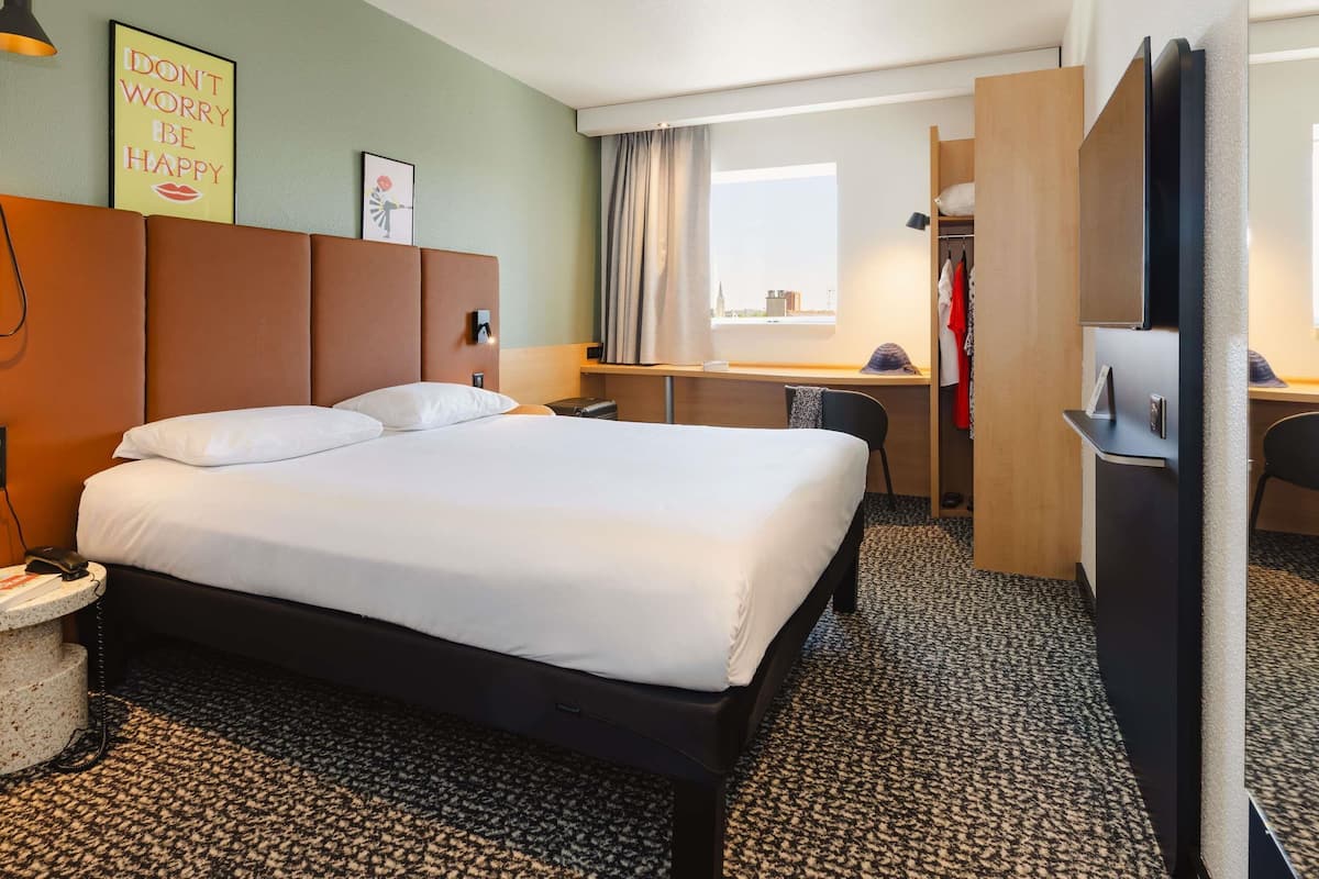 Quarto de hotel com uma cama grande, uma mesa, uma cadeira, um guarda-roupa e uma janela com cortinas.