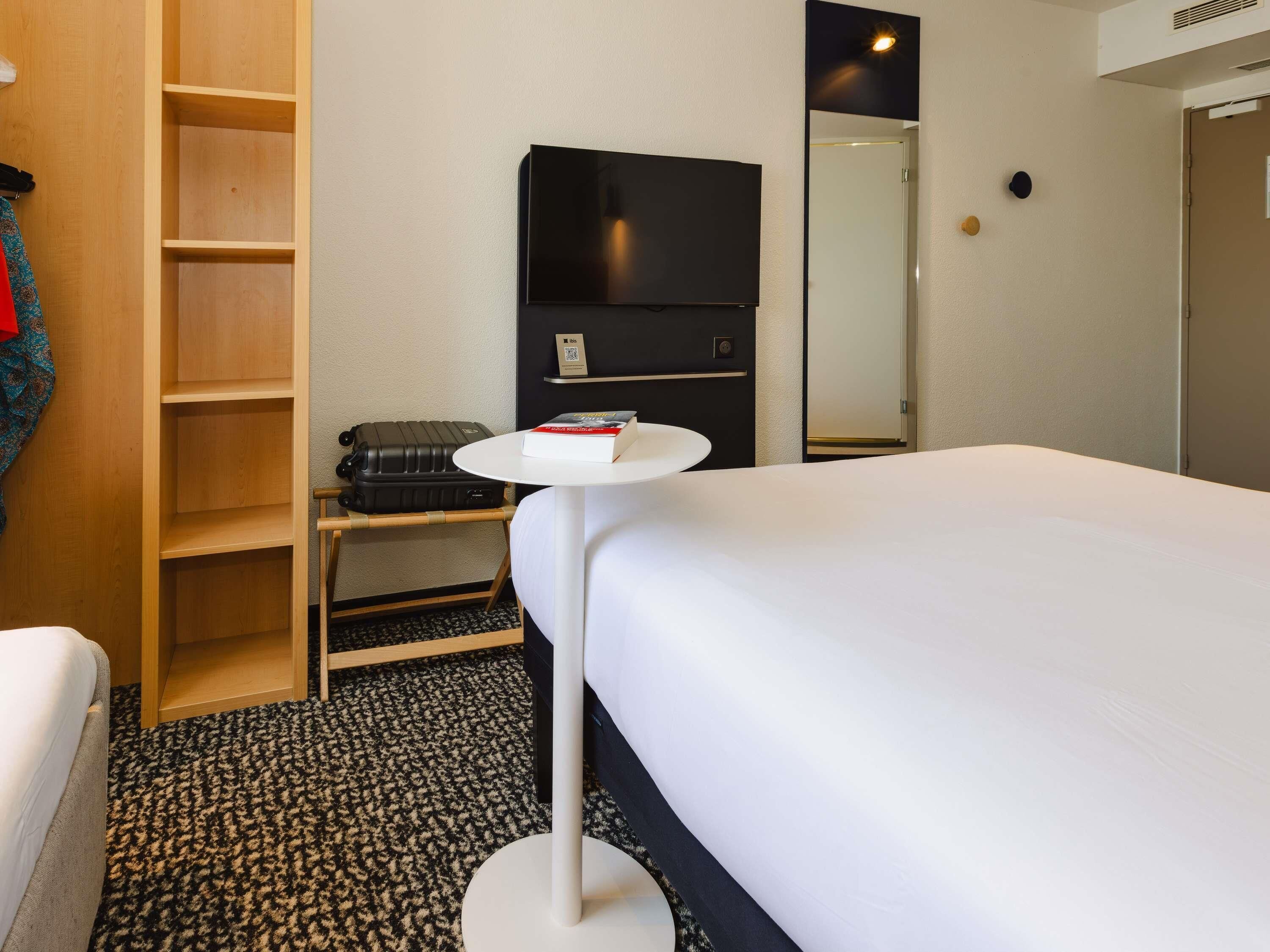 A hotel room with a bed, a small table with a book, a suitcase, a wardrobe, and a TV.