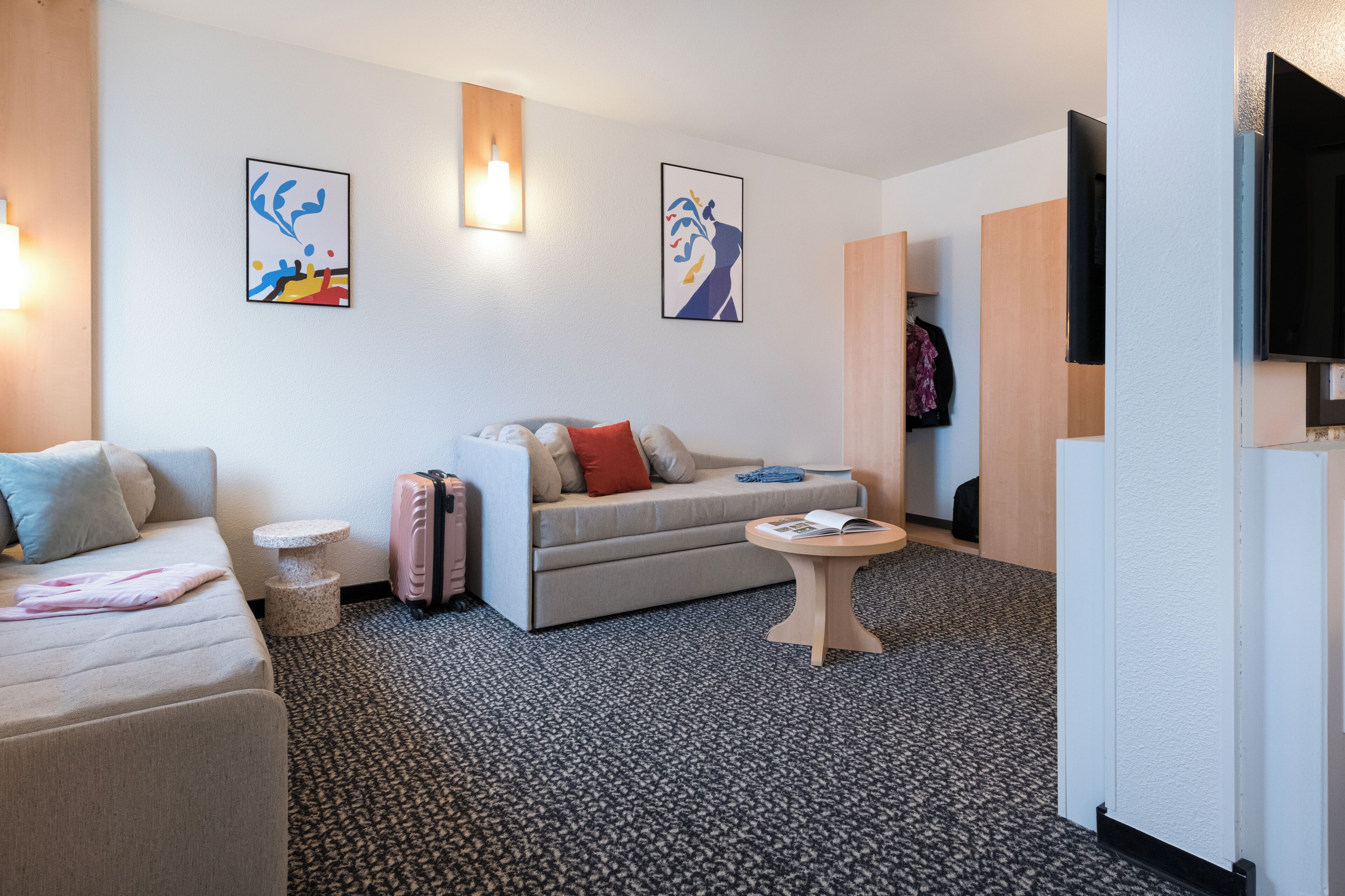A modern hotel room with a sofa, a small round table, and a bed. There are two artworks on the wall and a television on the right.