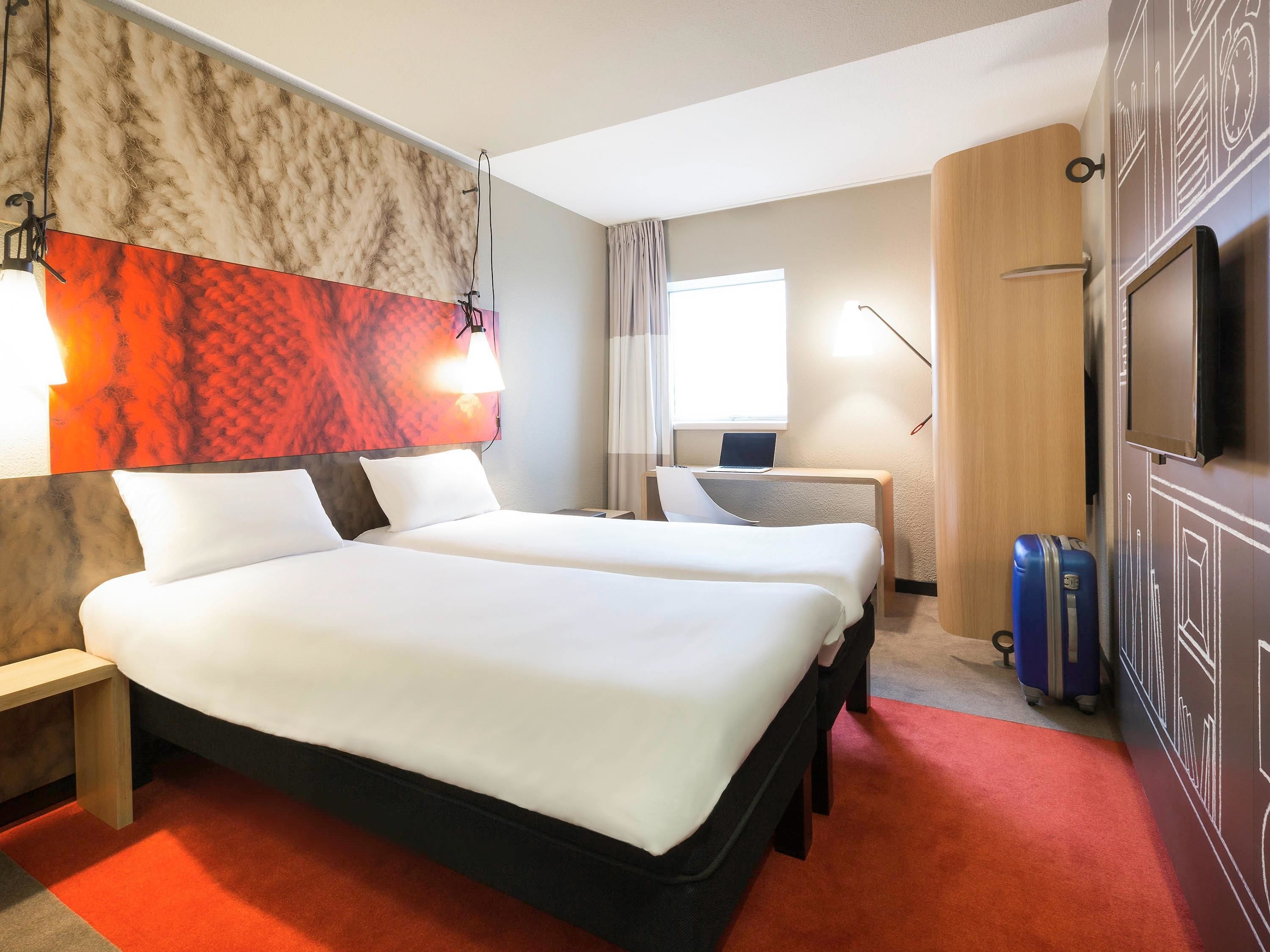 Ein Hotelzimmer mit zwei Betten, einem Schreibtisch, einem Laptop, einem Fernseher und einem Koffer.