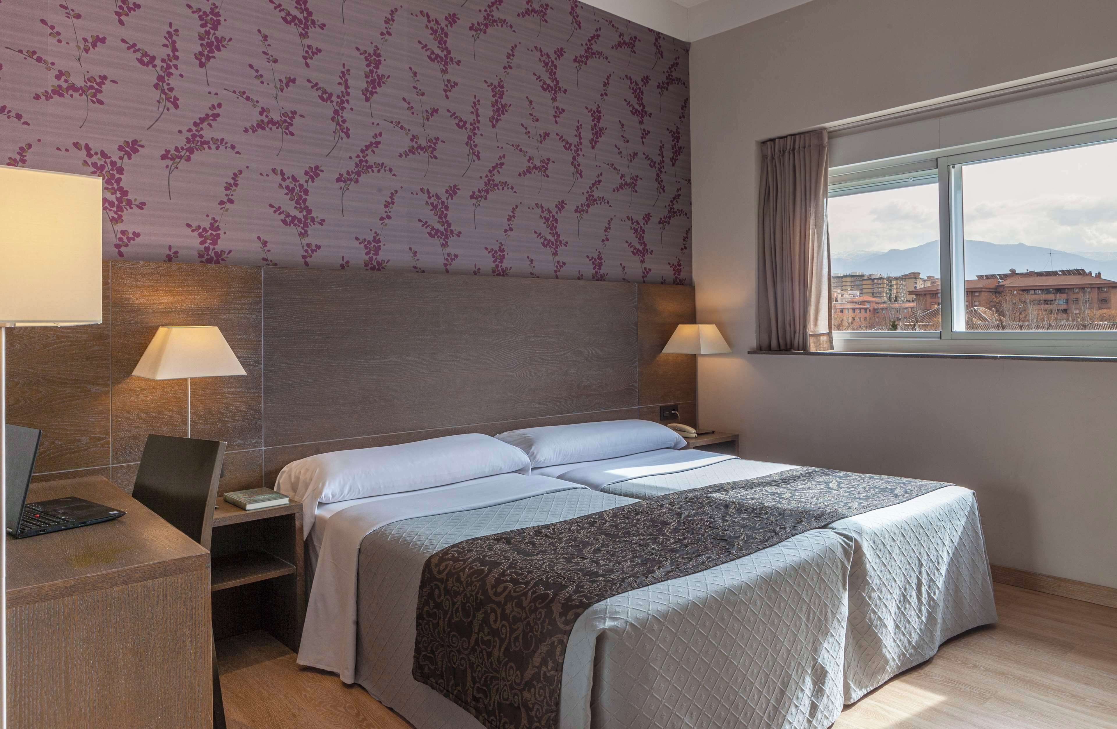 A hotel room with a bed, two bedside tables, a desk with a laptop, and a window with a view of buildings and mountains.
