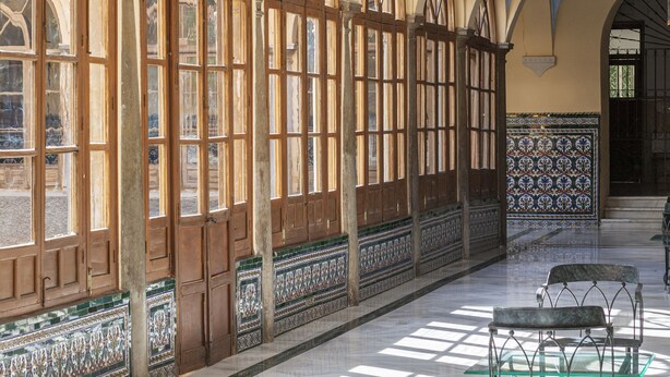 A spacious corridor with high arched windows and a tiled floor, leading to a room with a patterned wall.