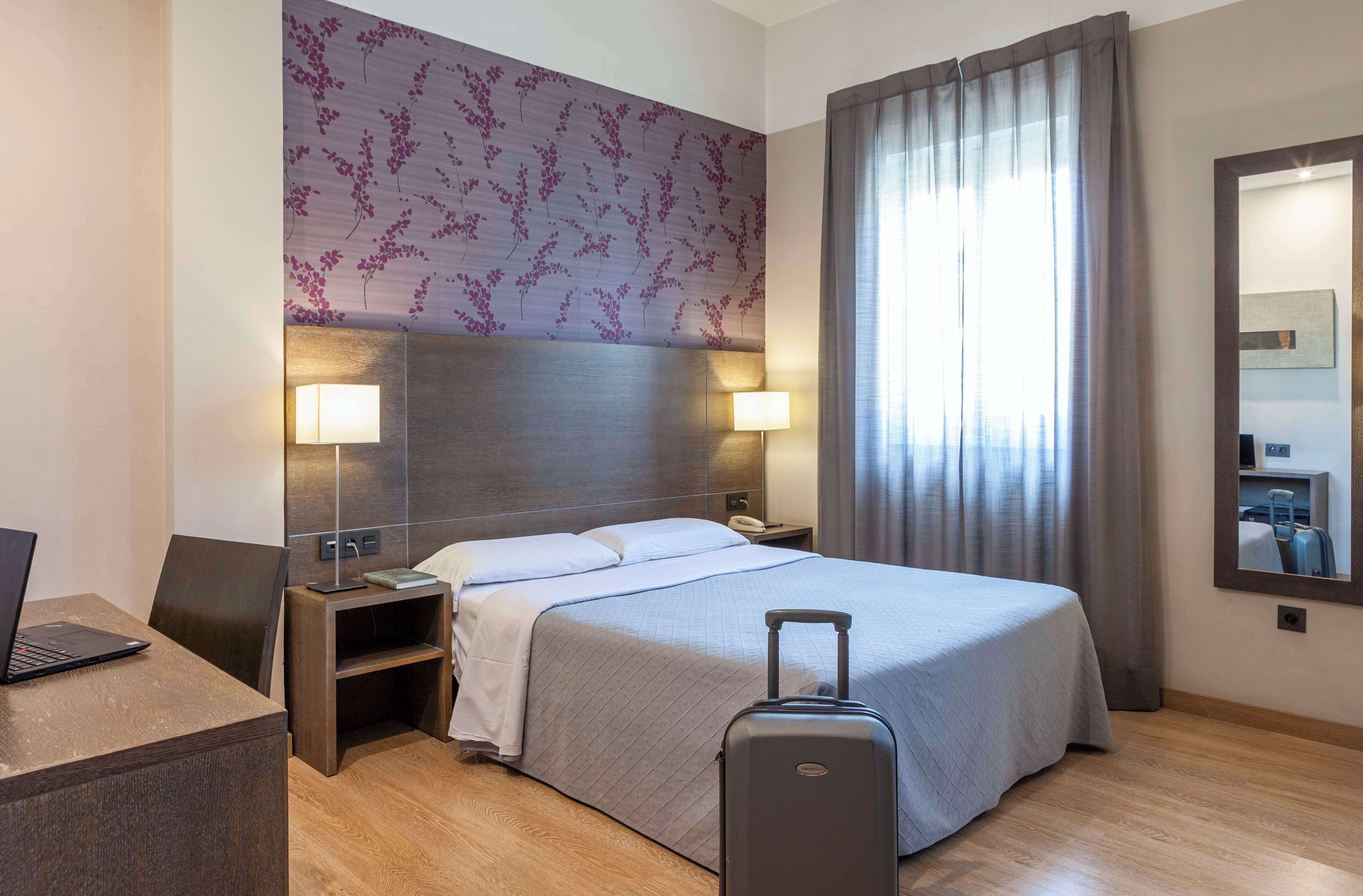 A hotel room with a bed, two bedside lamps, a desk with a laptop, a suitcase, and a large mirror on the wall.